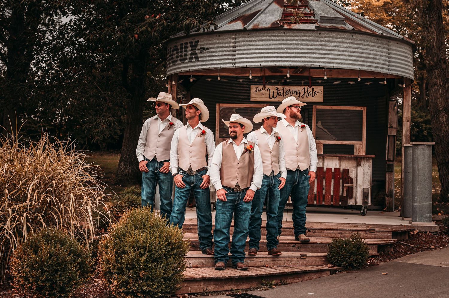 Five groomsmen in cowboy hats, white shirts, and jeans pose outside rustic venue building.