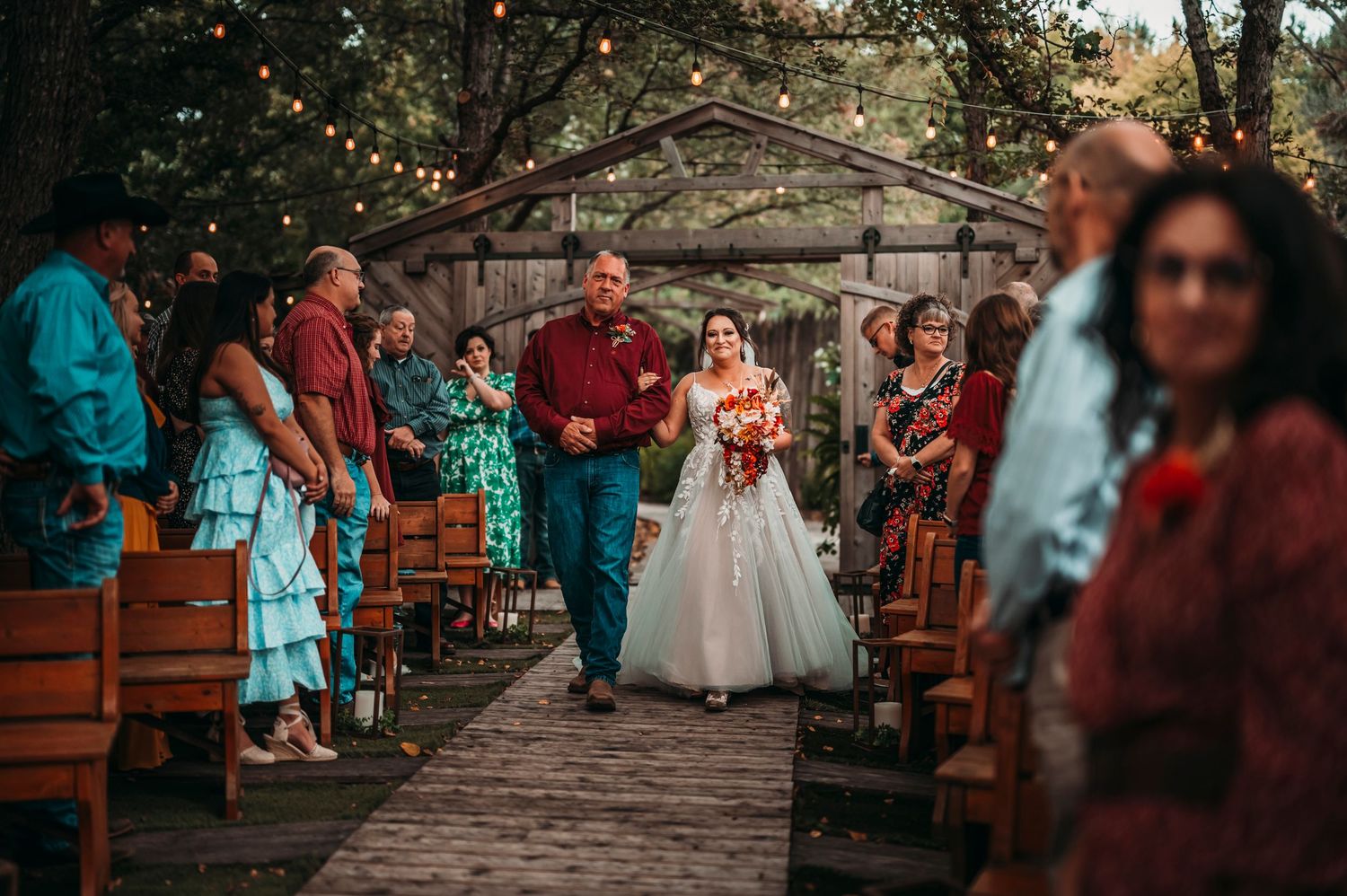Bride walks down outdoor aisle with escort past seated guests under string lights at dusk.