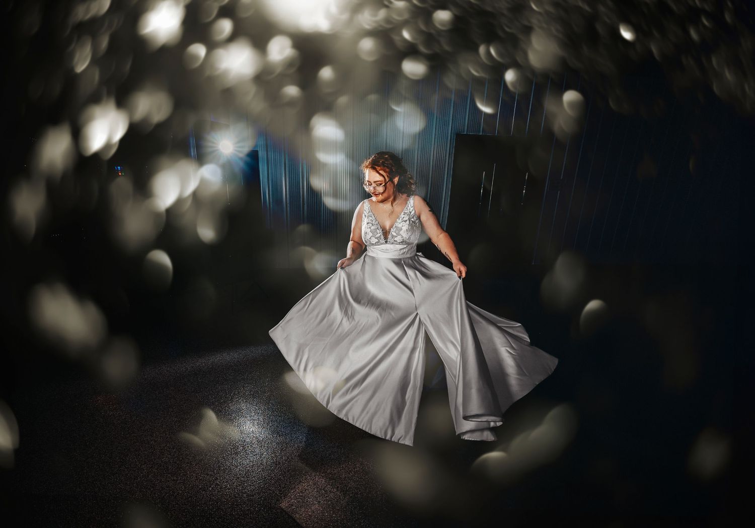 Woman in silver ball gown twirling elegantly in dramatic bokeh light at wedding reception.