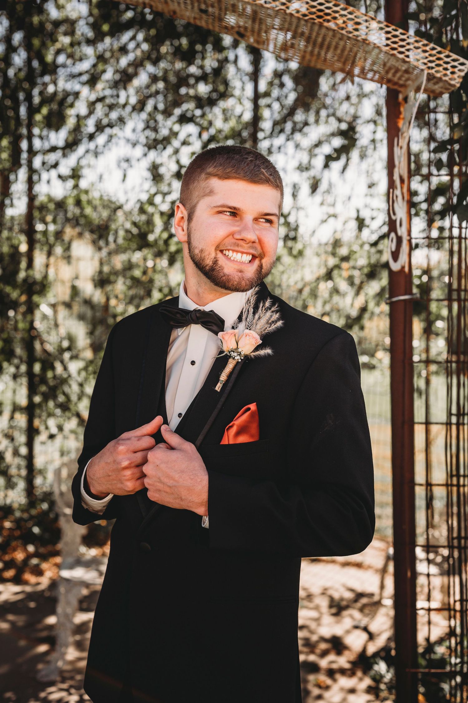 Smiling groom in black tuxedo with boutonniere and orange pocket square poses outdoors. Bessie's Barn