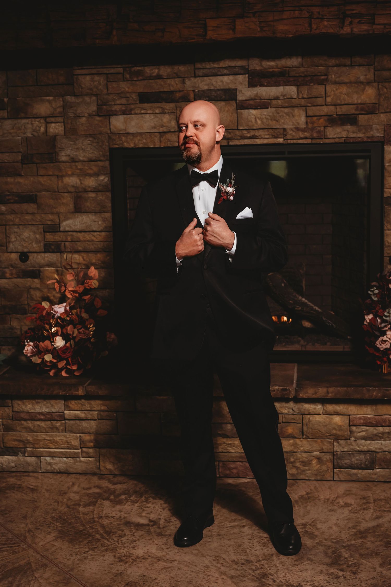Dapper groom in black tuxedo poses confidently before stone fireplace with floral arrangements.