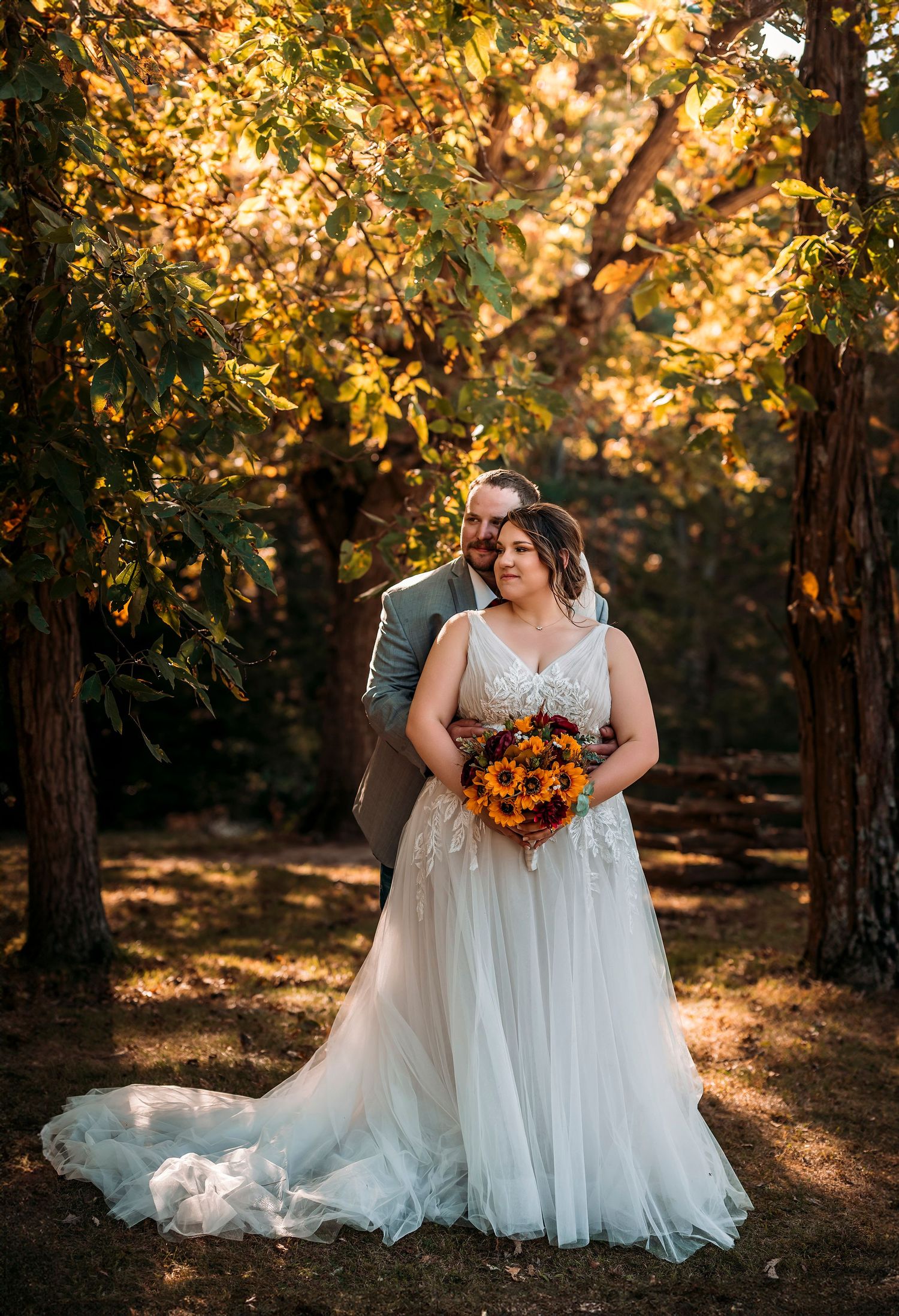 Bride and groom pose romantically among golden autumn trees, bride holding sunflower bouquet in elegant white gown.