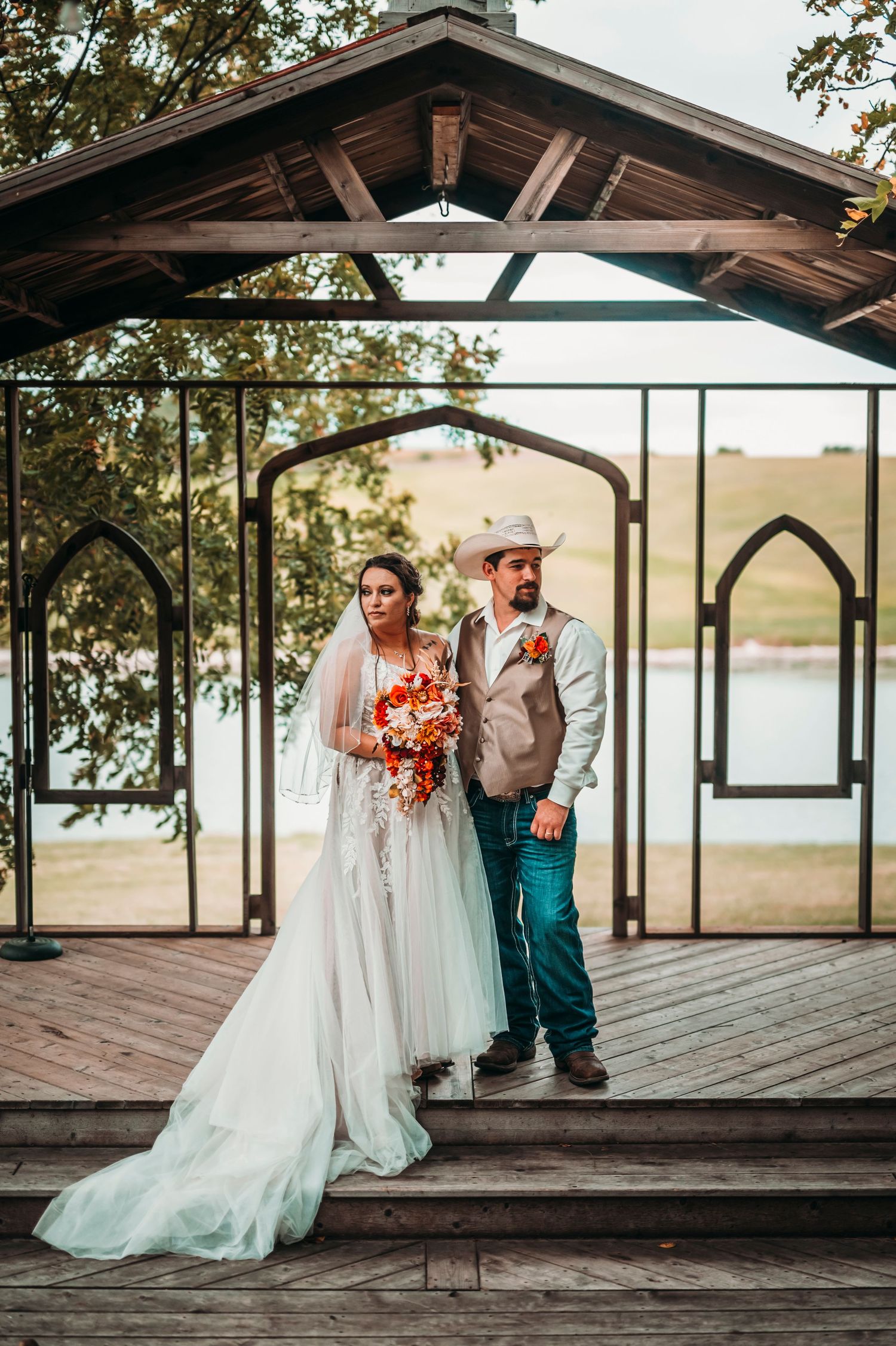 Bride and groom stand under rustic wooden pergola with lake backdrop, groom in cowboy hat.