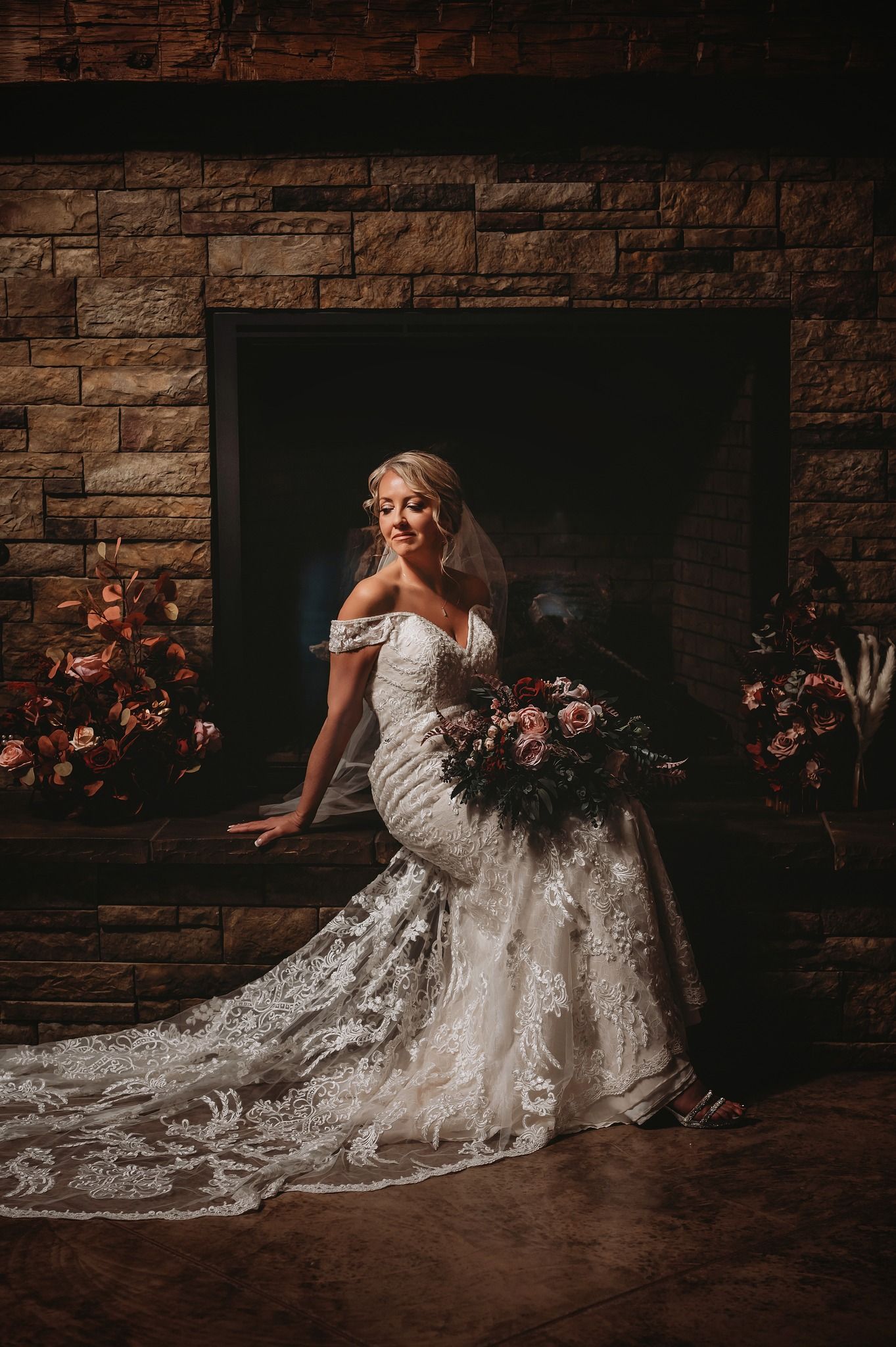 Stunning bride in lace mermaid gown sits by stone fireplace holding dramatic burgundy bouquet.