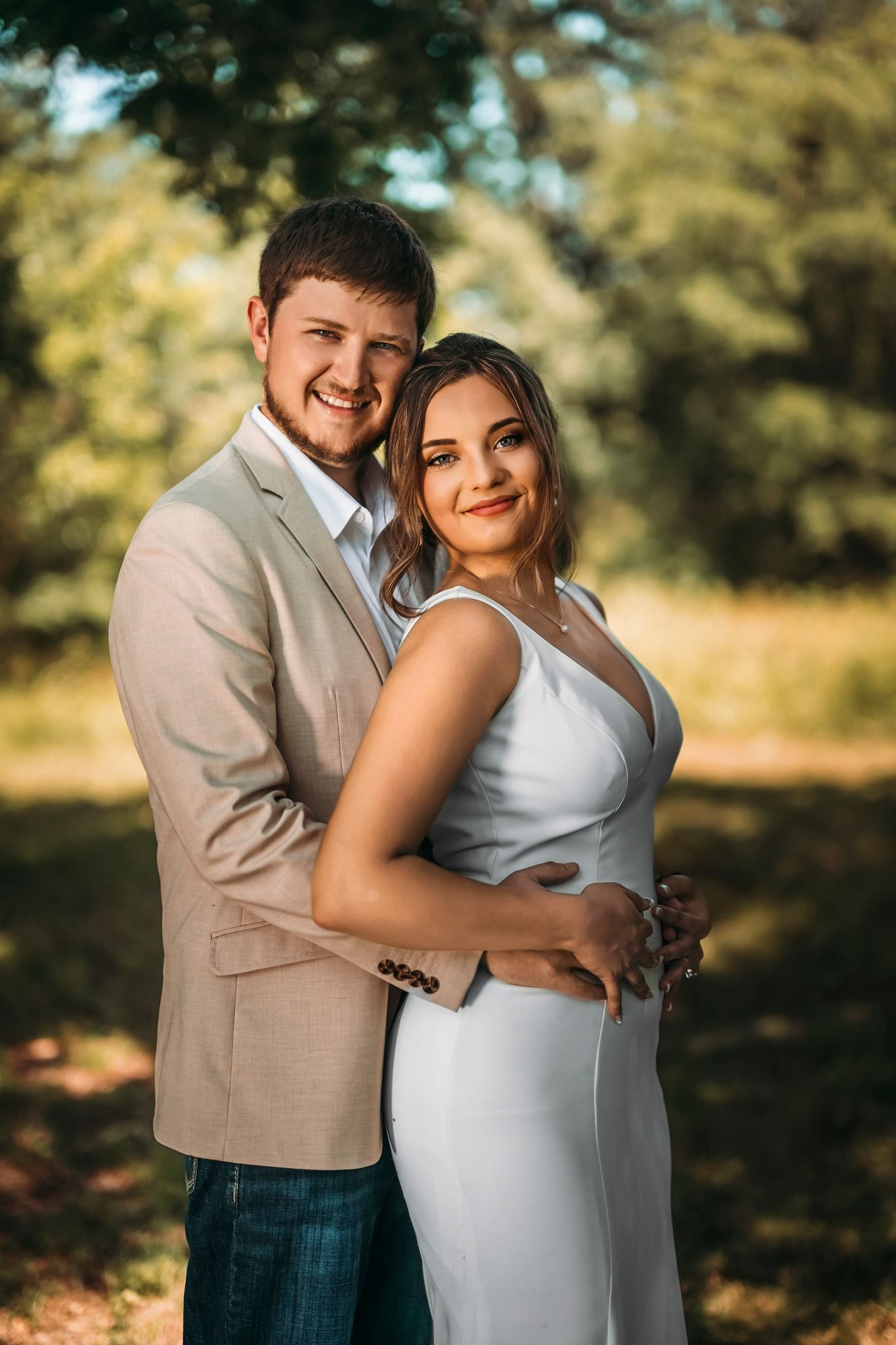 Couple embraces and smiles together in sunlit park, bride in white dress, groom in tan blazer.