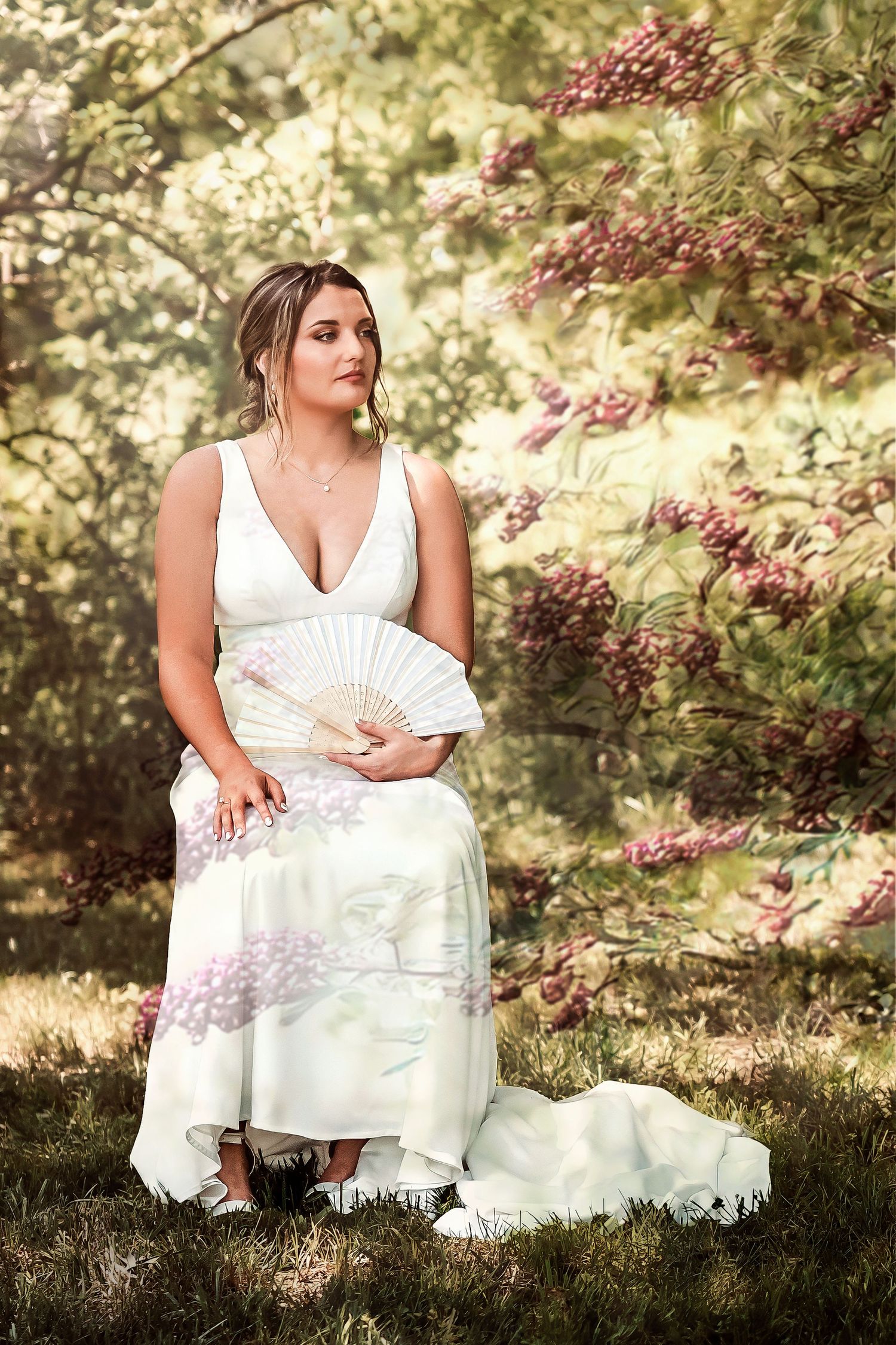 Bride in white dress holding floral fan seated near blooming pink flowering trees outdoors.