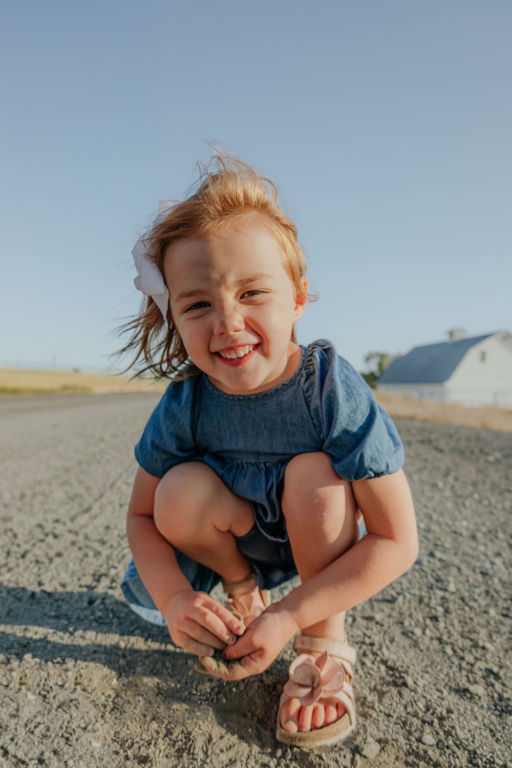 Child in denim dress crouching and smiling on rocky ground during golden hour.
