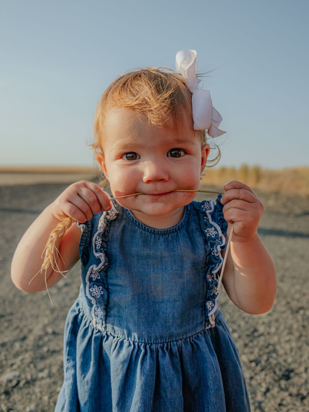 Young child in denim dress with white bow smiling at camera.