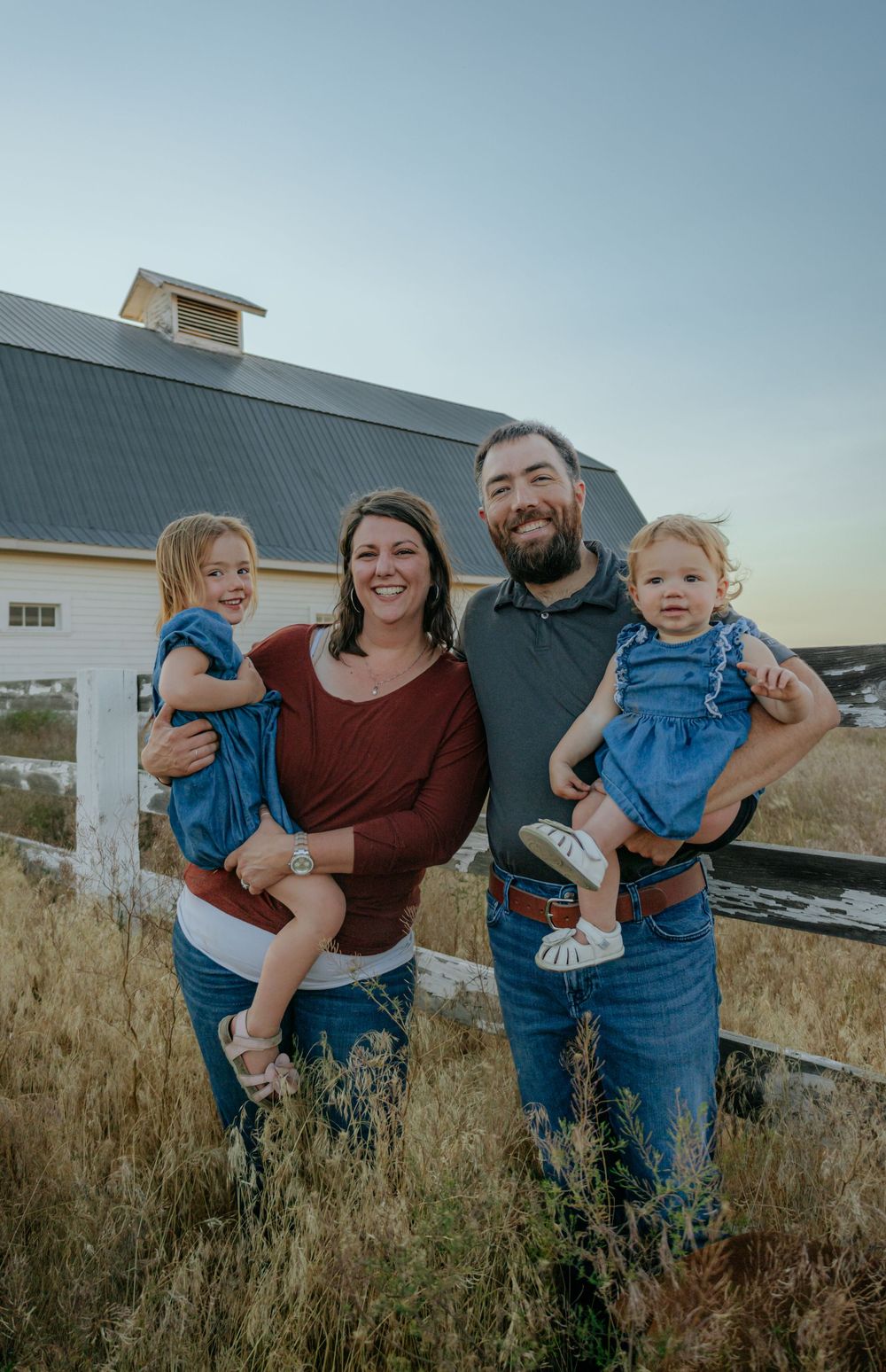 Family plays together in a grassy field at sunset near a white barn.