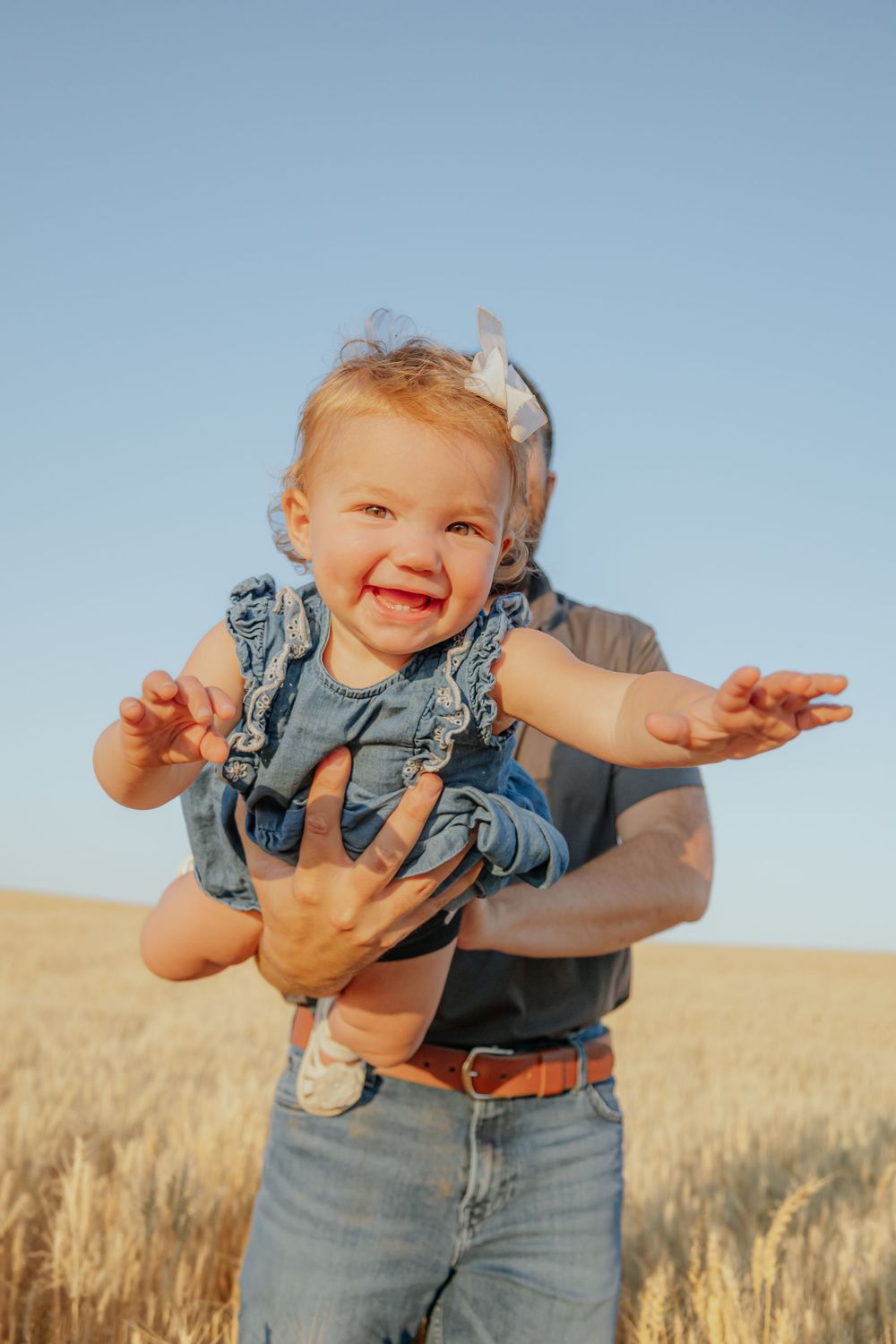 Baby in denim overalls smiles and plays while being held outdoors in a wheat field.