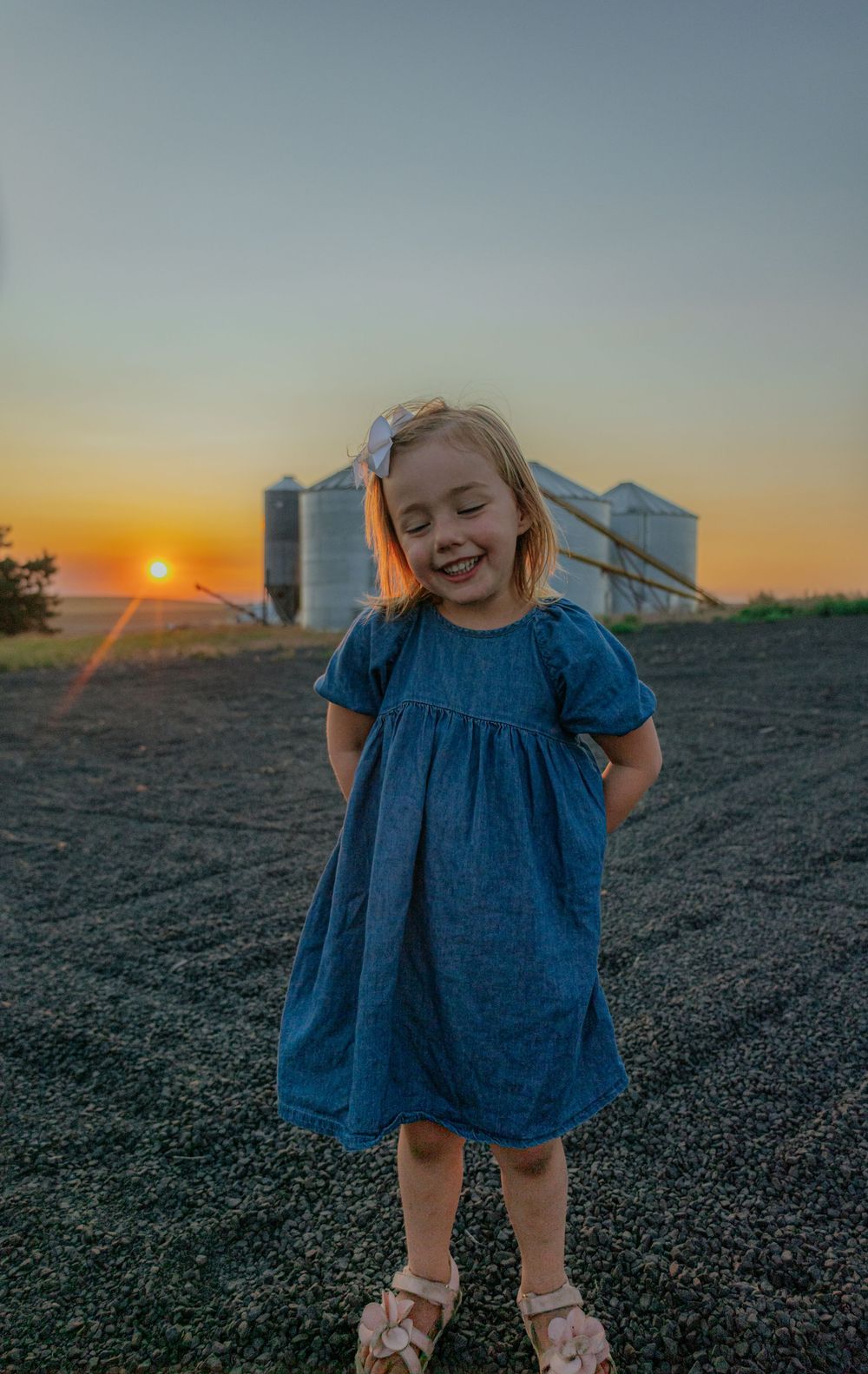 A child in a blue dress dances playfully in a field at sunset with farm buildings in the background.