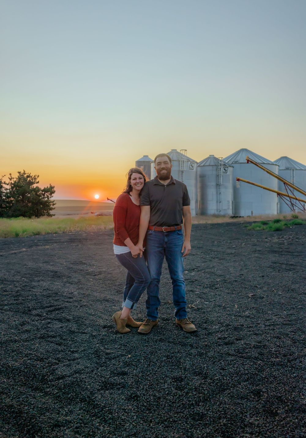 Couple poses for photos at sunset with grain silos in background.