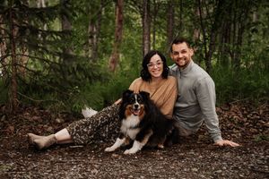 A couple sits with their Australian Shepherd dog on a forest trail surrounded by evergreen trees.