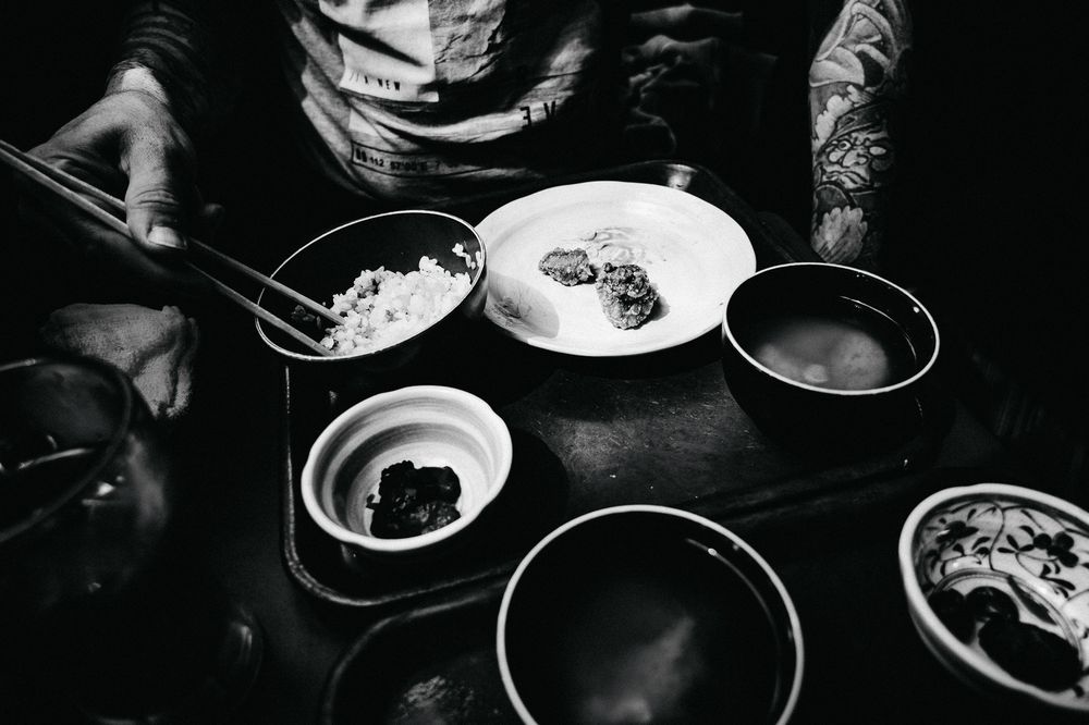 Black and white photograph shows multiple plates of food and drinks on a dark table in a high contrast style.