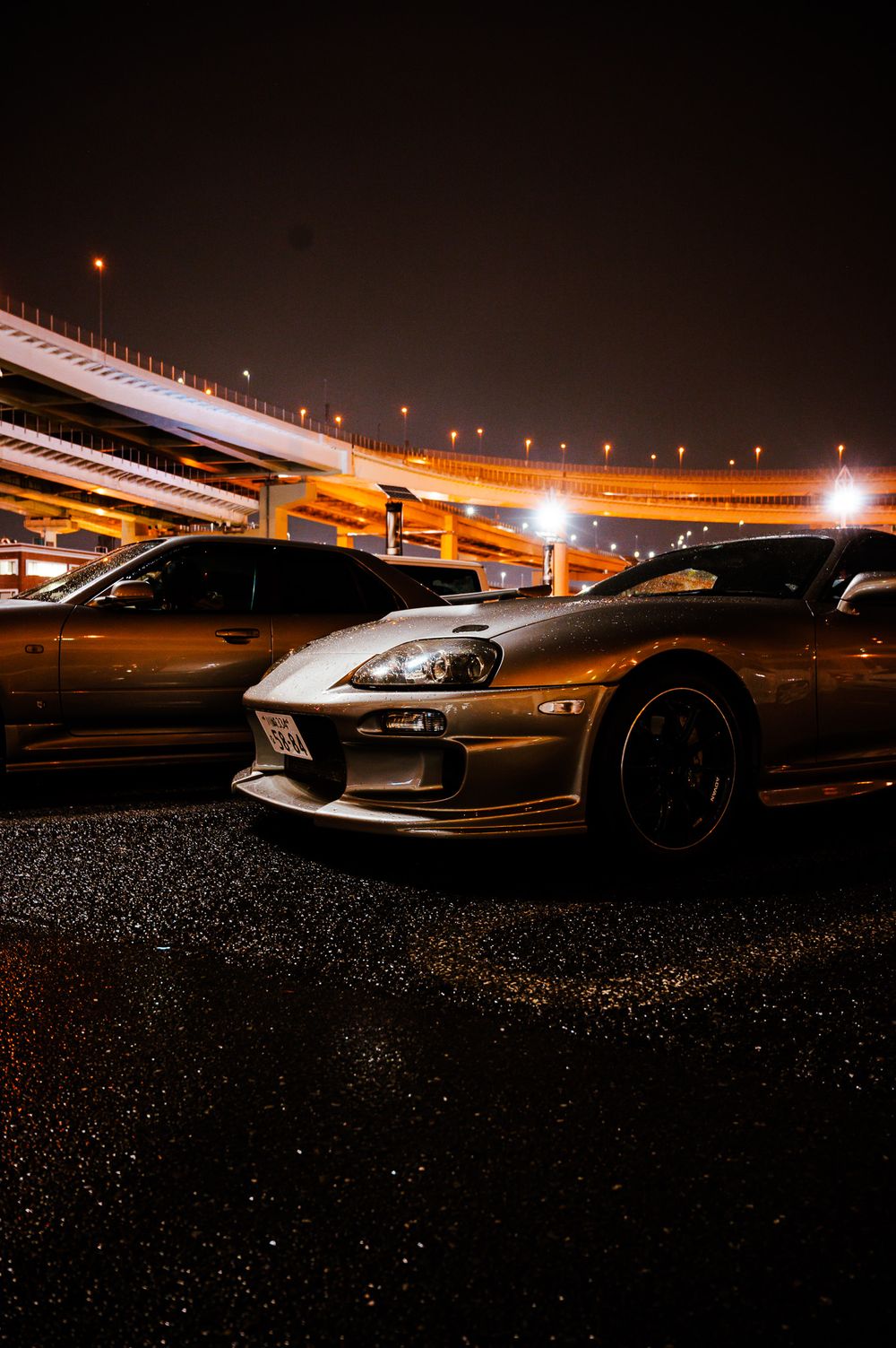 Sleek sports cars parked at night under illuminated highway overpasses creating dramatic lighting.