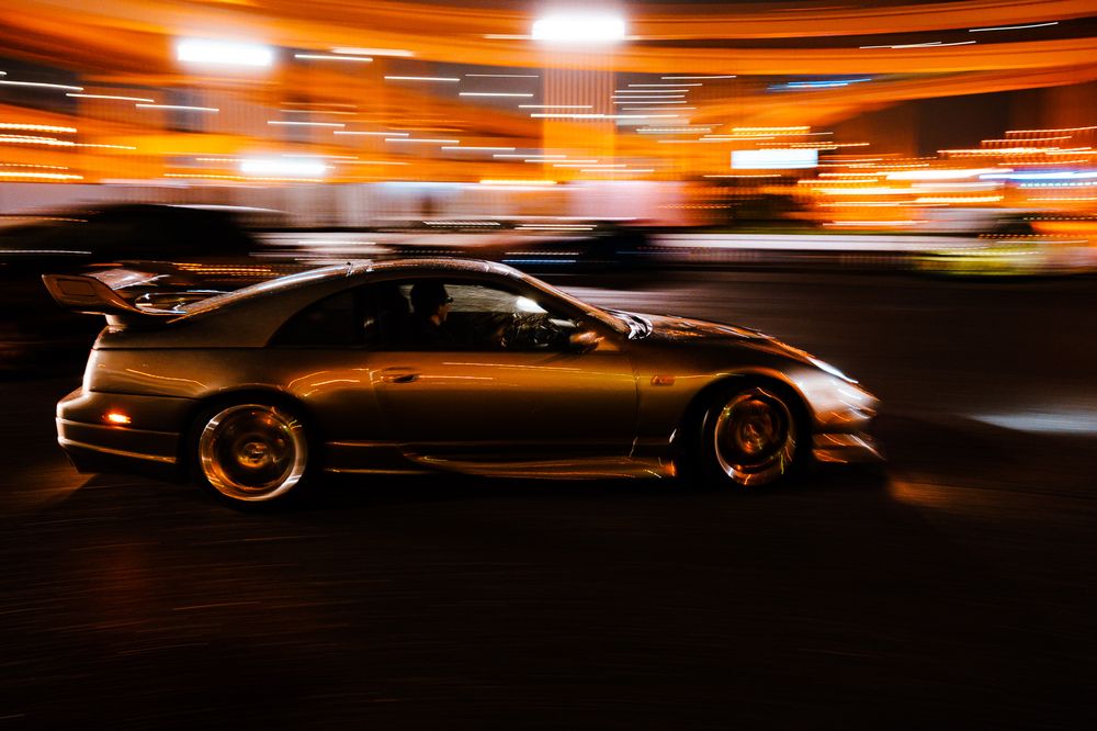 Long exposure shot of race cars creating illuminated light trails against an orange-lit racing track at night.