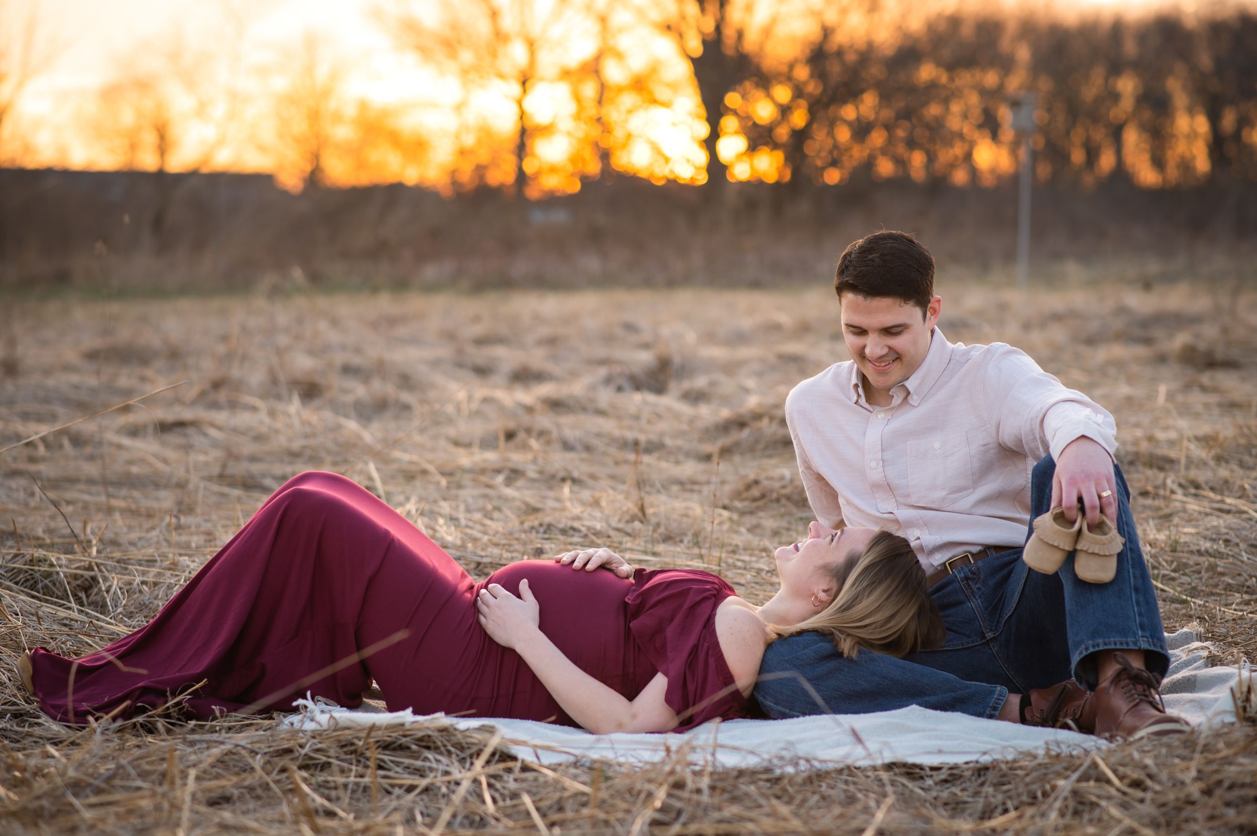 A couple dressed in white and burgundy sitting on a blanket in a golden field at sunset.