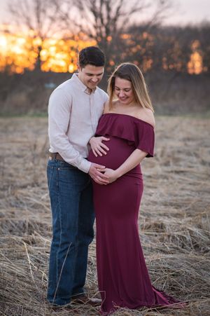 Couple shares tender moment in rural field at sunset as they celebrate impending arrival in burgundy maternity dress.