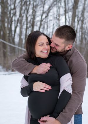 A cozy winter maternity photoshoot with a couple embracing in the snow-covered woods.