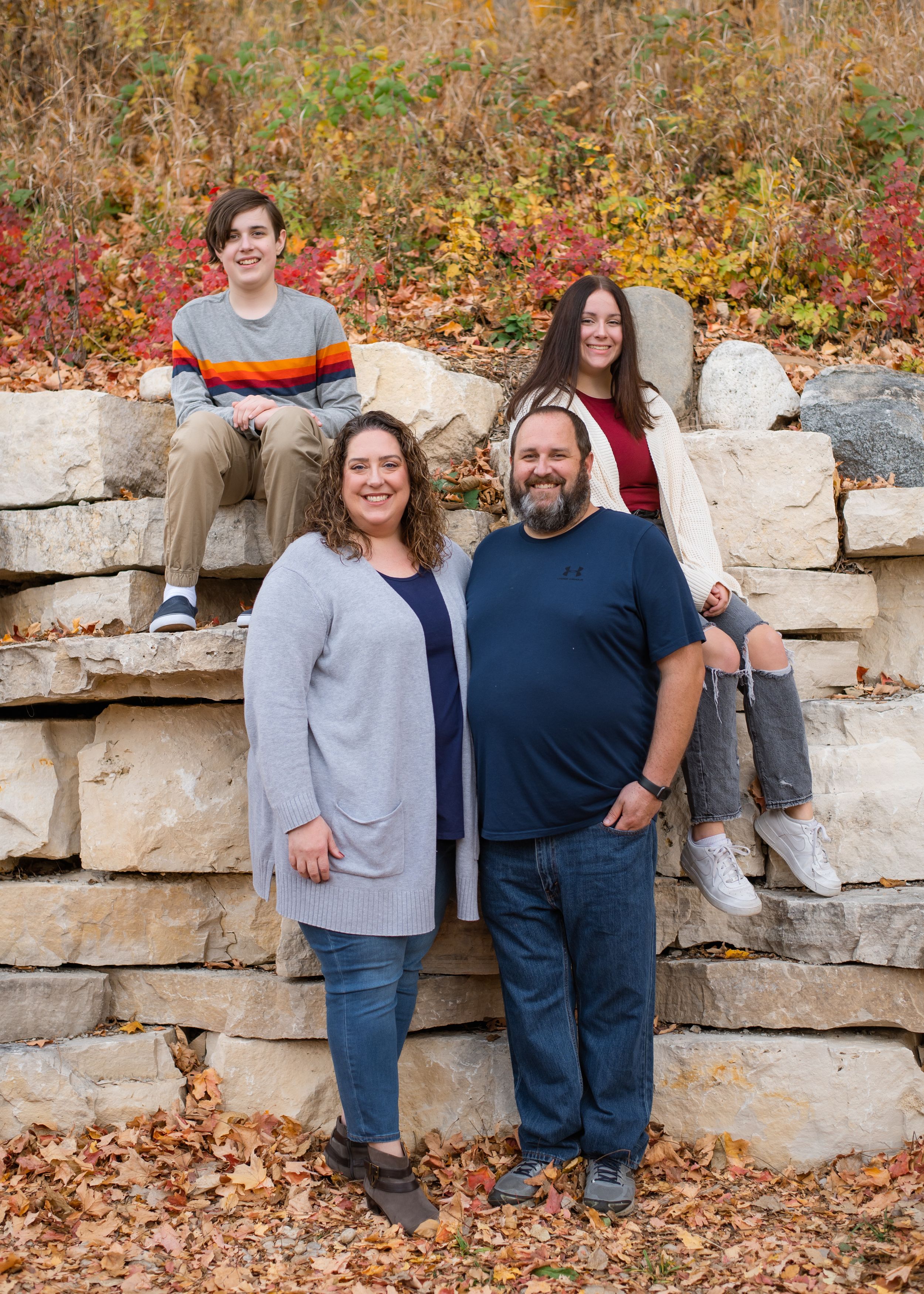 Family members sit together on stone wall surrounded by autumn leaves in casual attire.
