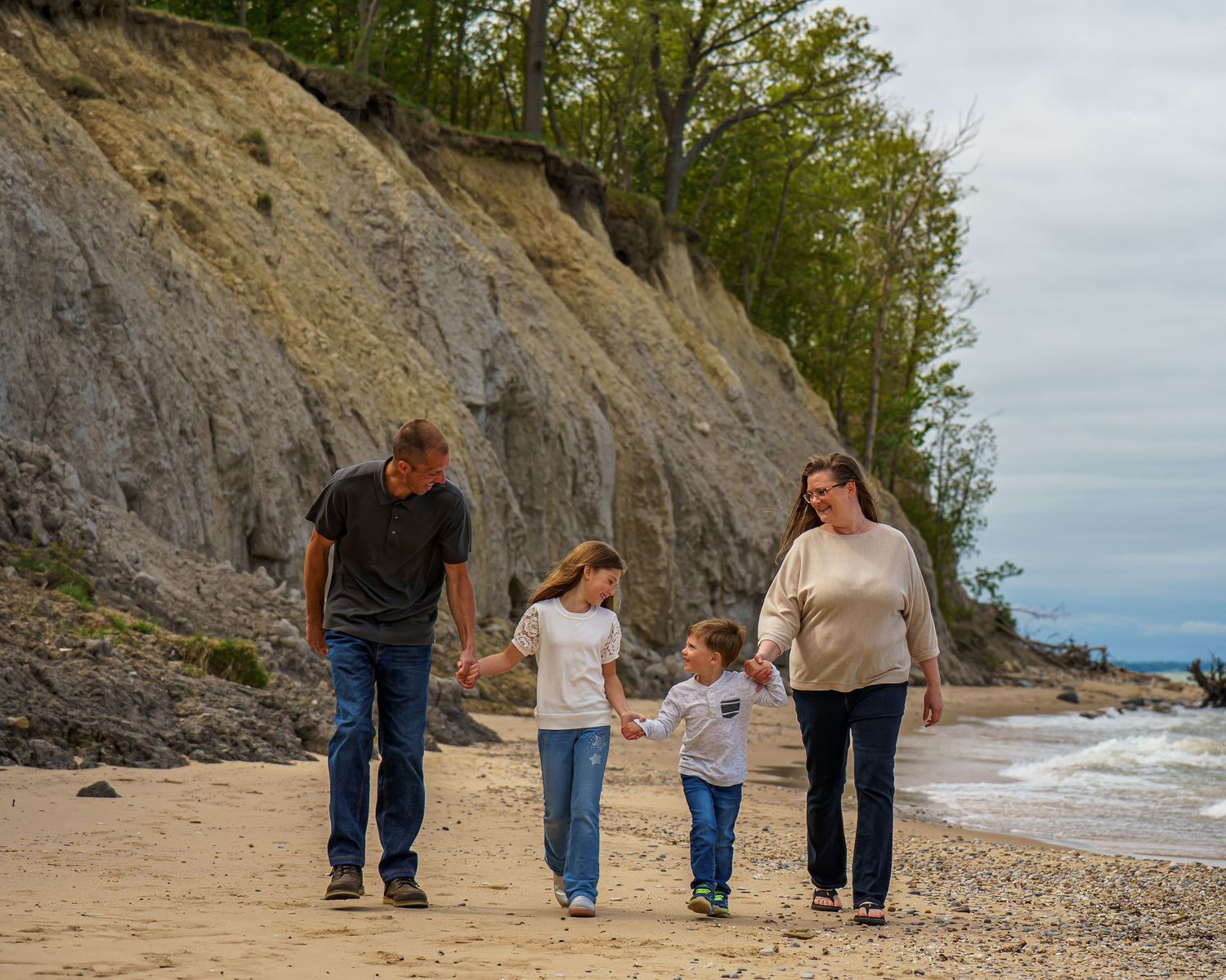 Family of four walking hand-in-hand along a sandy beach with scenic bluffs and trees in the background.