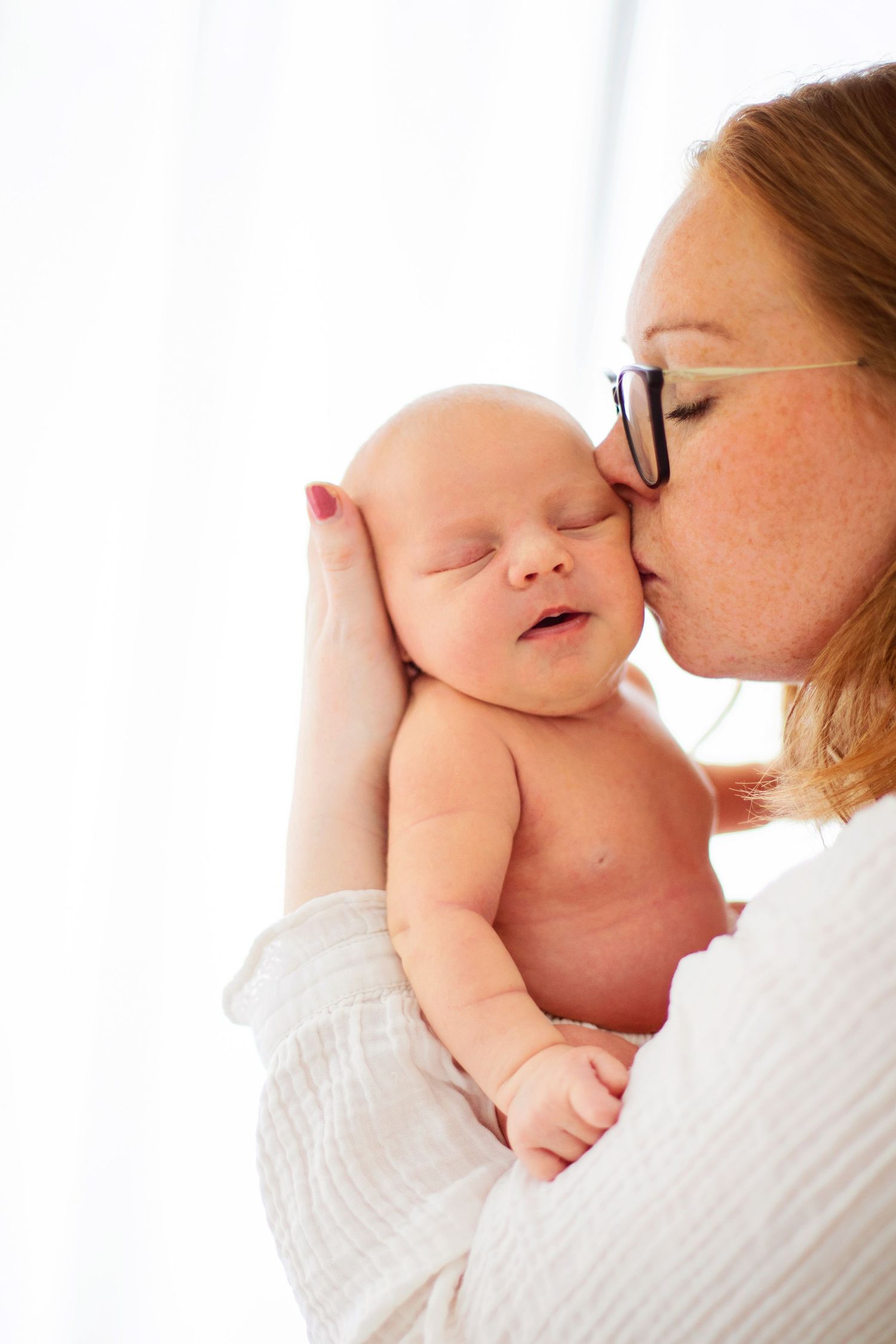 A freckled redhead mother with glasses tenderly kisses her sleeping newborn baby on the cheek.