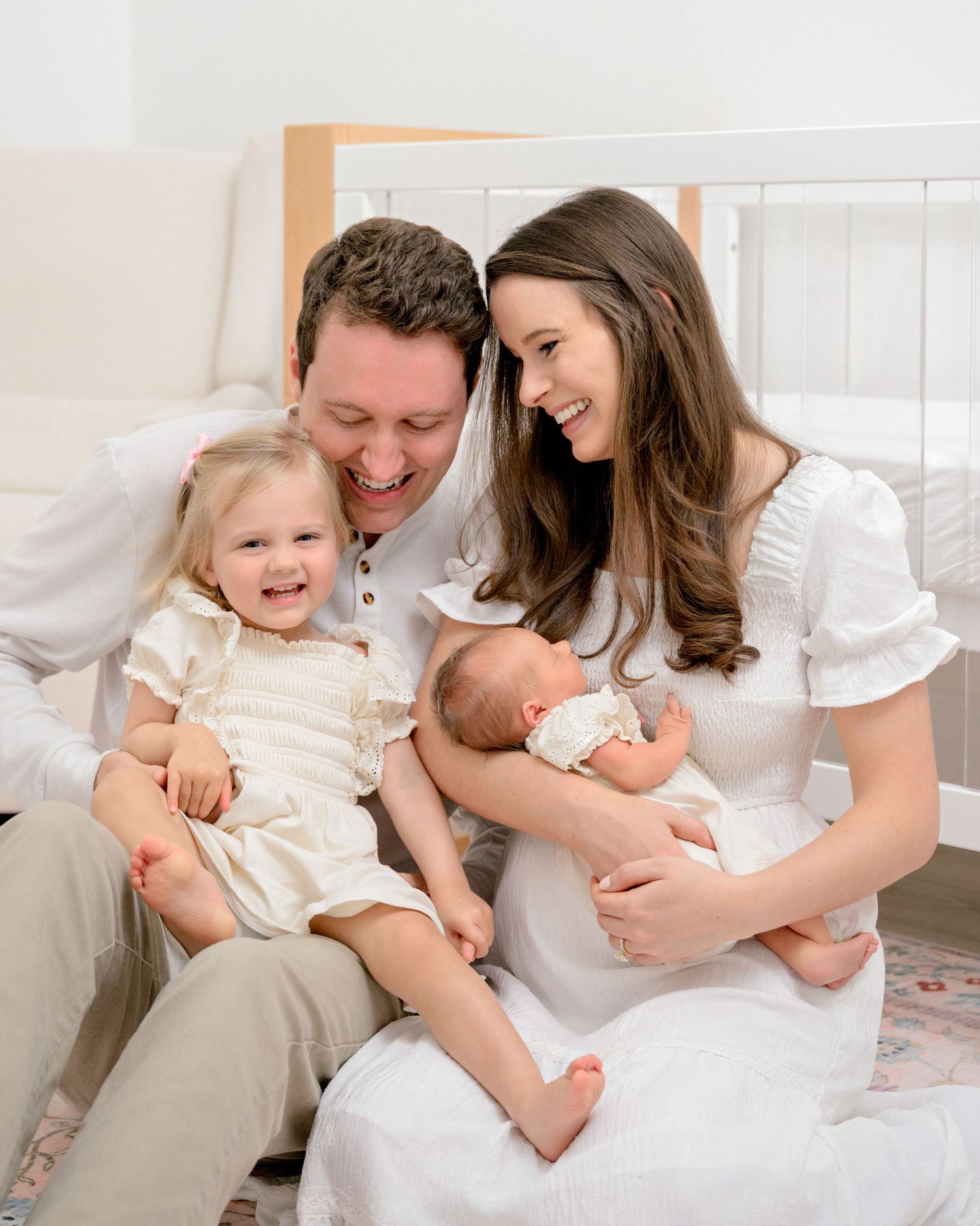 Happy family of four in white outfits, parents smiling with toddler girl and newborn baby during professional photo session.