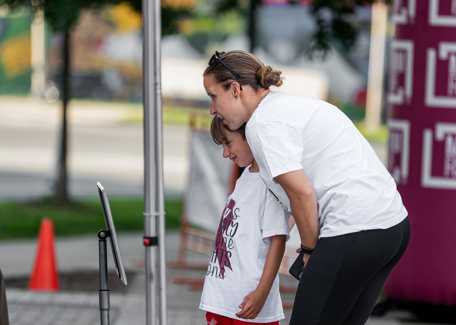 A woman in a white event shirt bends down to speak with a young child at an outdoor community gathering.