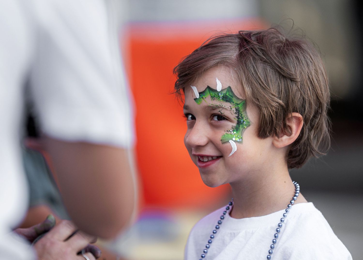 Young boy with a green dinosaur face paint design smiling at an outdoor festival event.