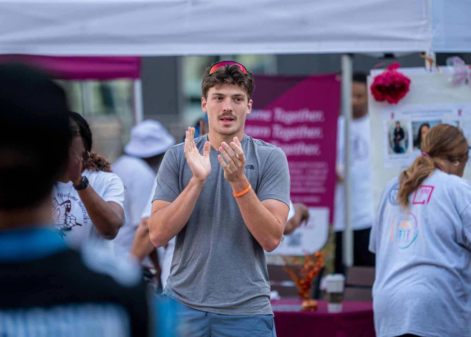 Man clapping and engaging with crowd at an outdoor charity event near a purple and white tent.