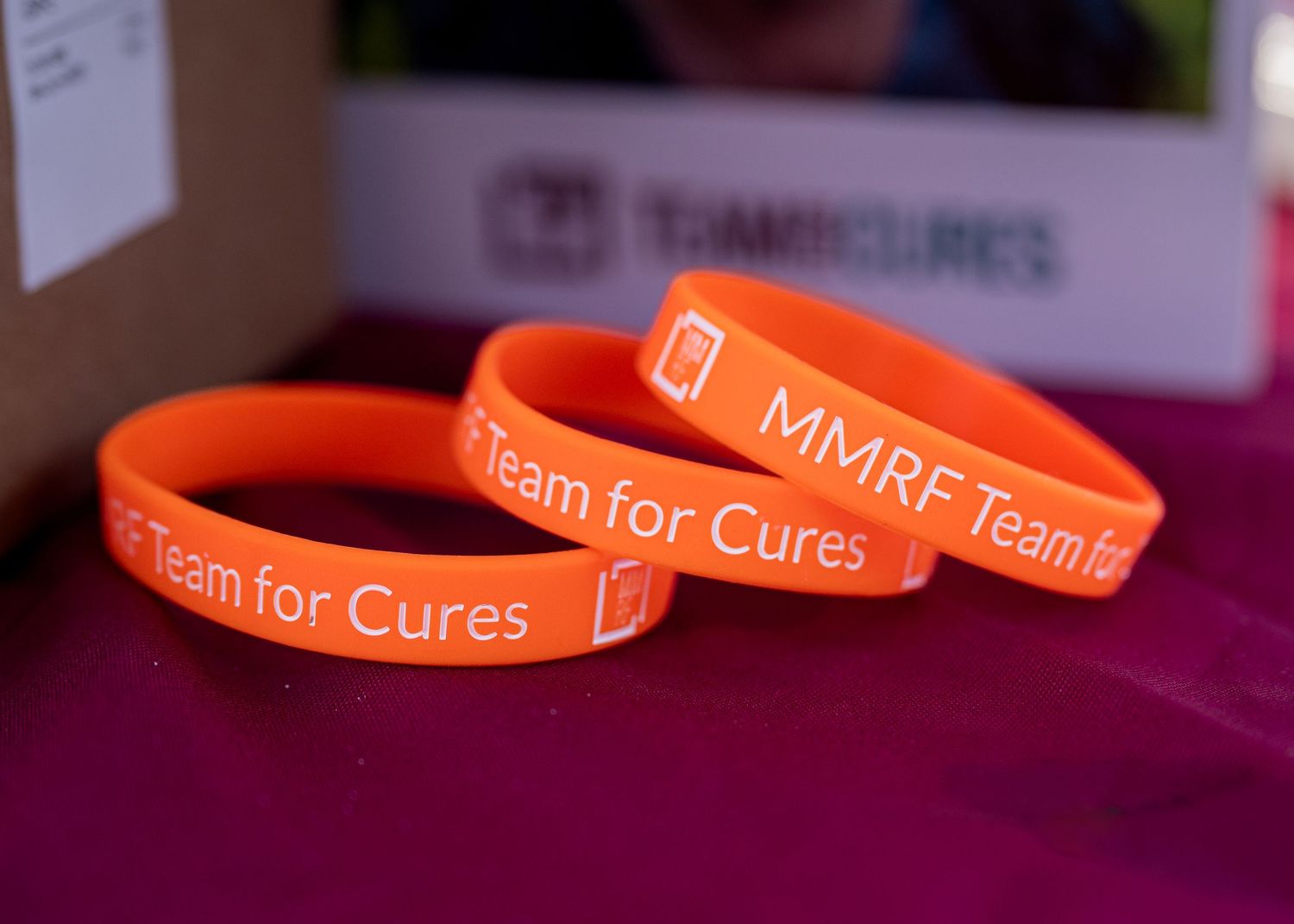 Three orange MMRF Team for Cures silicone charity wristbands displayed on a purple surface.