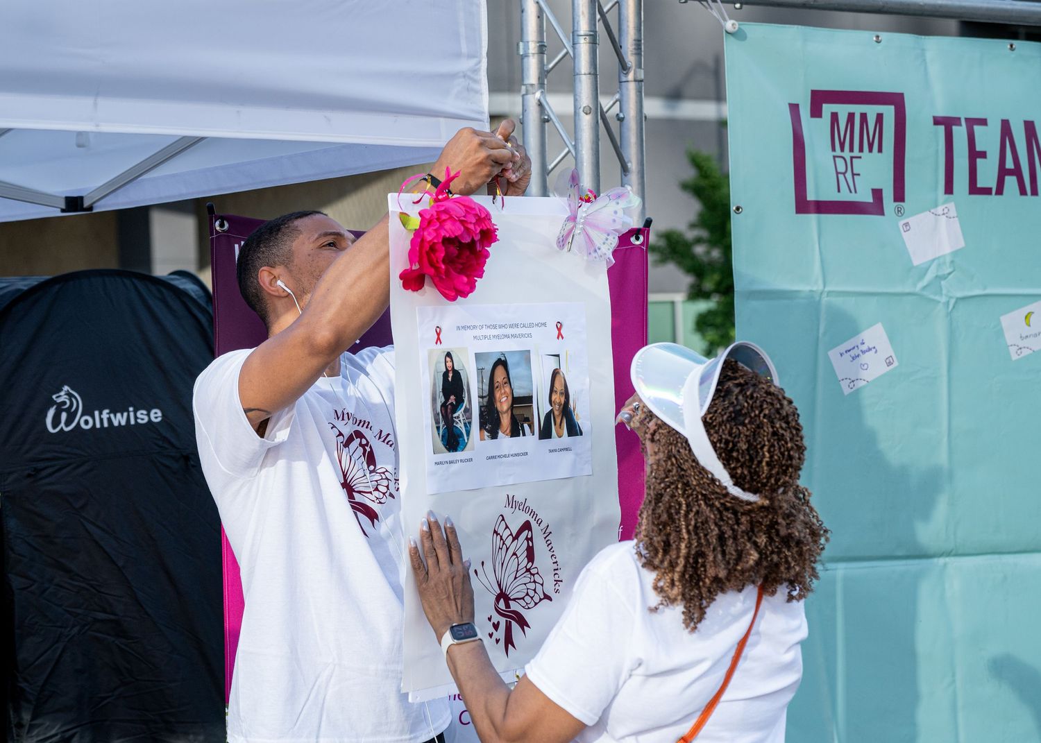 Two people pin photos and a pink flower to a display board at a Multiple Myeloma Research Foundation event.