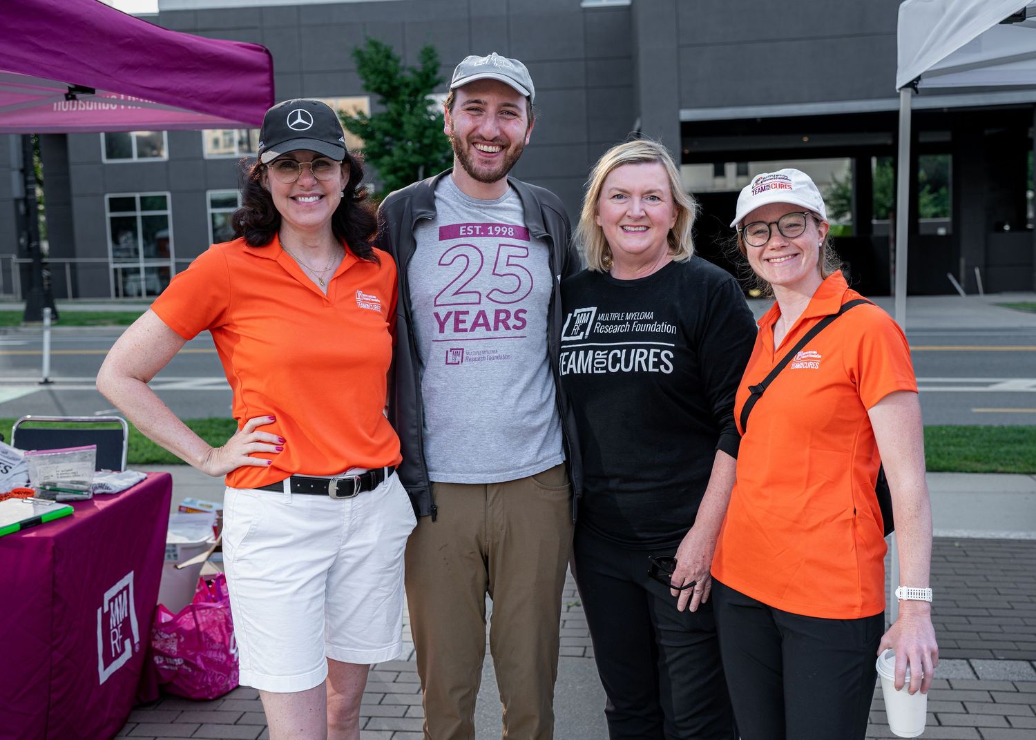Four people smiling together at a 25-year anniversary fundraising event outdoors, wearing orange branded shirts.