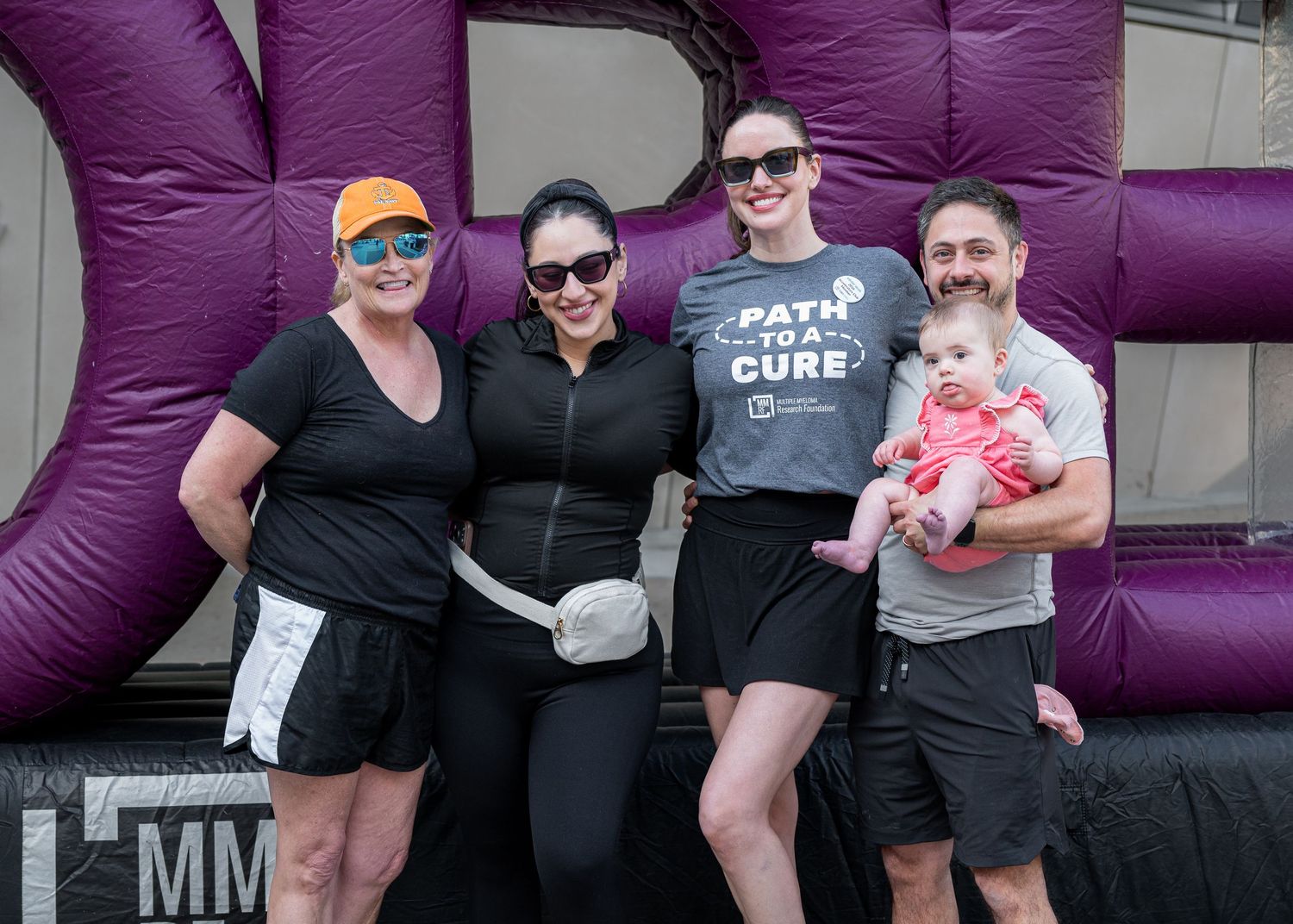 Group of four adults and a baby posing together at a charity walk event in front of a purple inflatable.