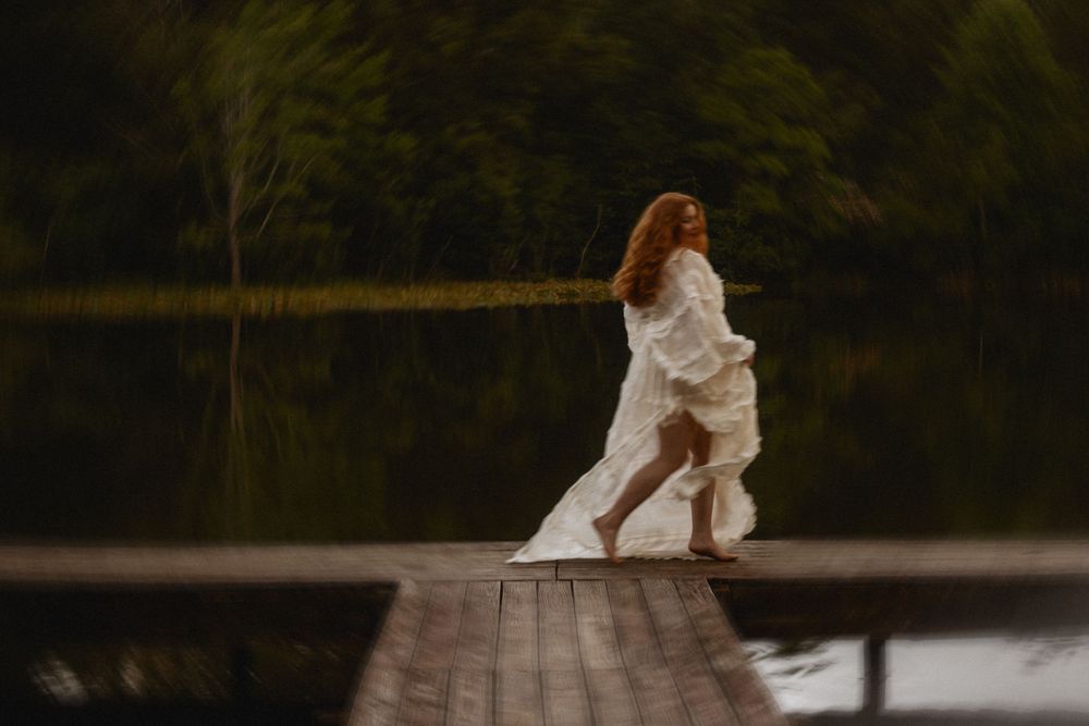 Woman in flowing white dress dancing on wooden dock by a serene lake surrounded by lush green trees.