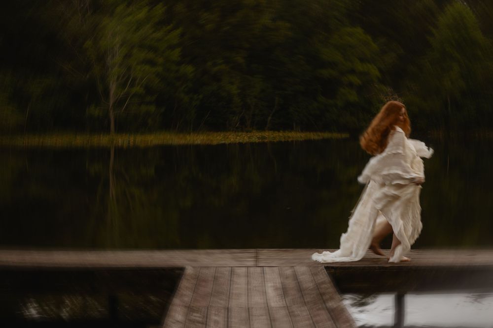 A red-haired woman in a flowing white dress stands on a wooden dock overlooking a calm, tree-lined lake.