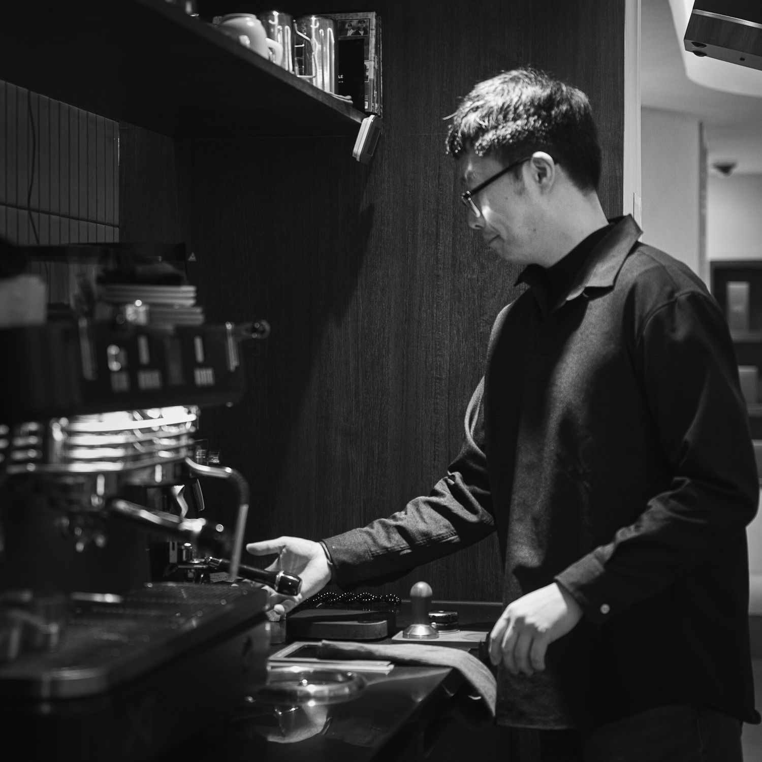 Black and white photo of a man wearing glasses working at a coffee espresso machine in a cafe.