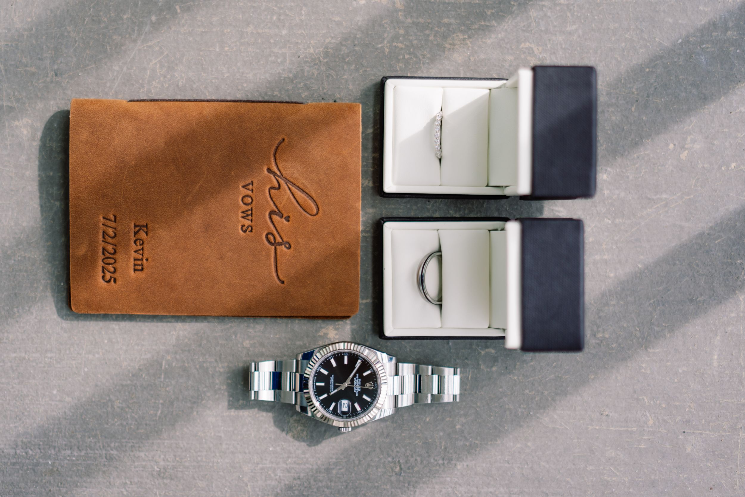Luxury stainless steel watch displayed alongside two ring boxes and a personalized brown leather wallet on a gray surface.