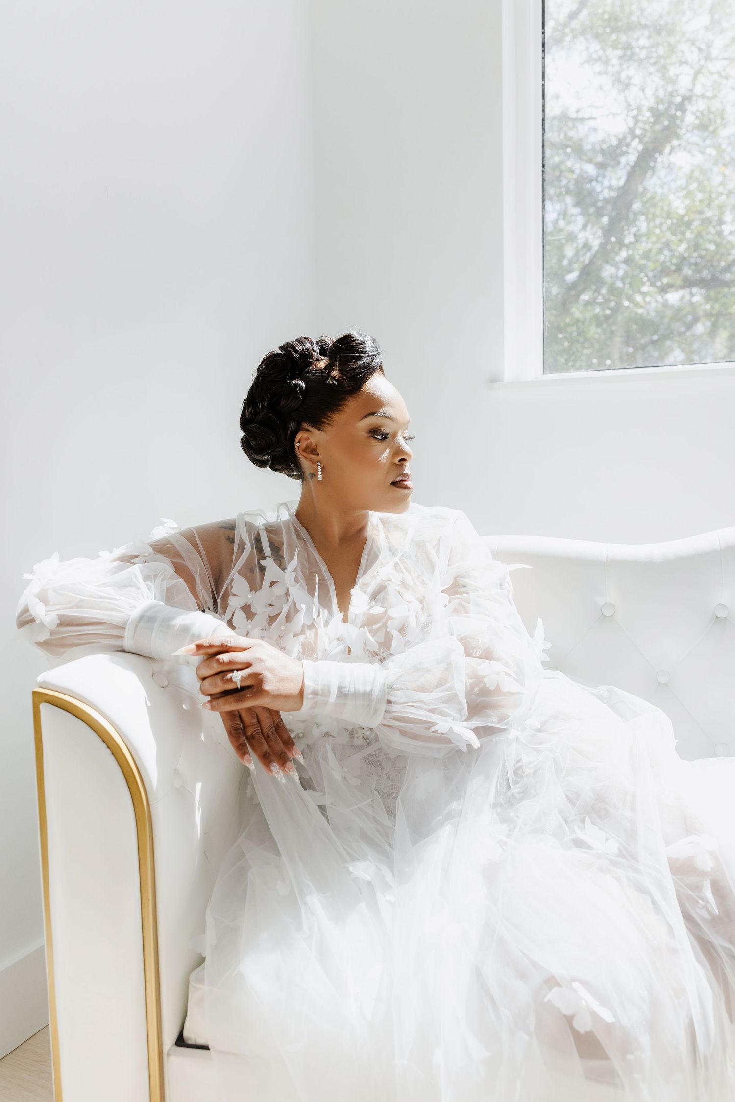 Bride in white sheer butterfly robe sitting on a white tufted chair, gazing out a bright window.