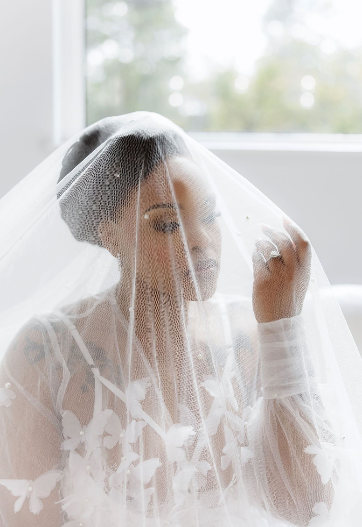 Tattooed bride in butterfly veil gazing down in soft natural light during bridal portrait session.