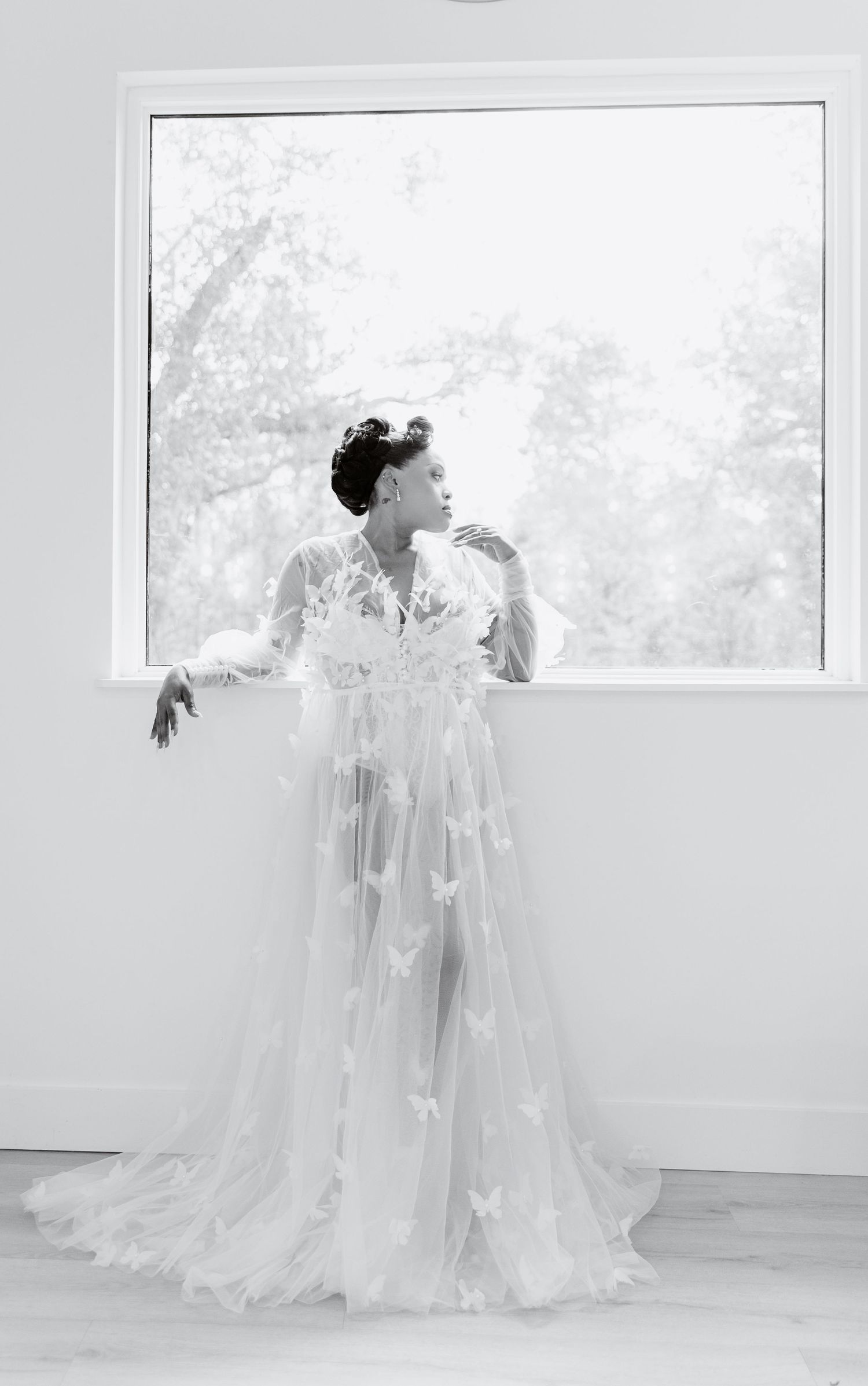 Black and white portrait of bride in sheer butterfly tulle robe standing elegantly before a large window.