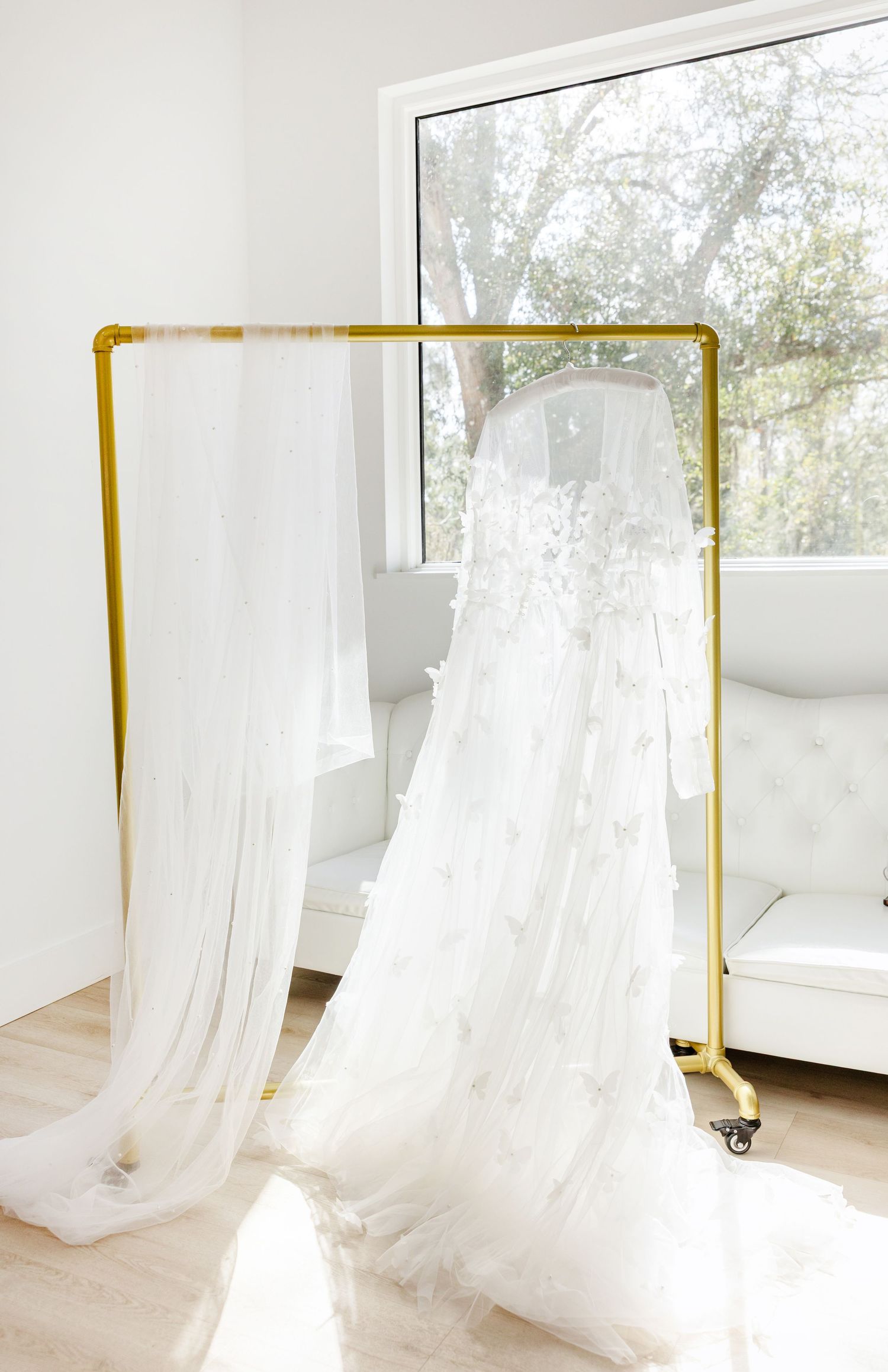White floral applique wedding gown with veil displayed on a gold garment rack in a bright white room.