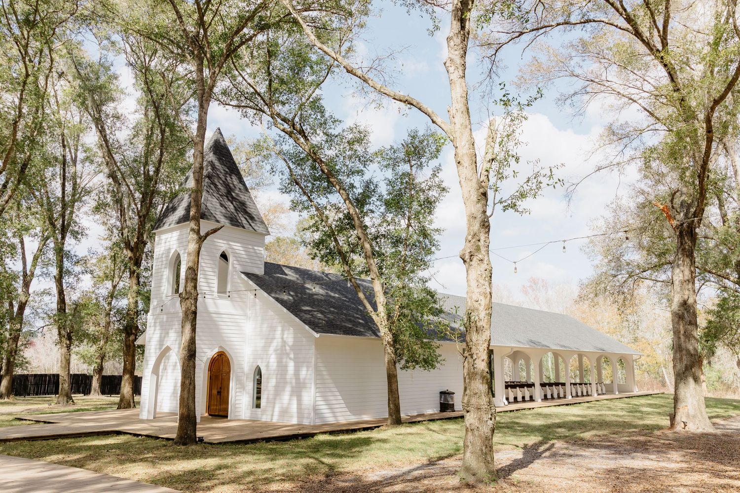 White chapel wedding venue with steeple surrounded by tall trees and adjacent reception tent.