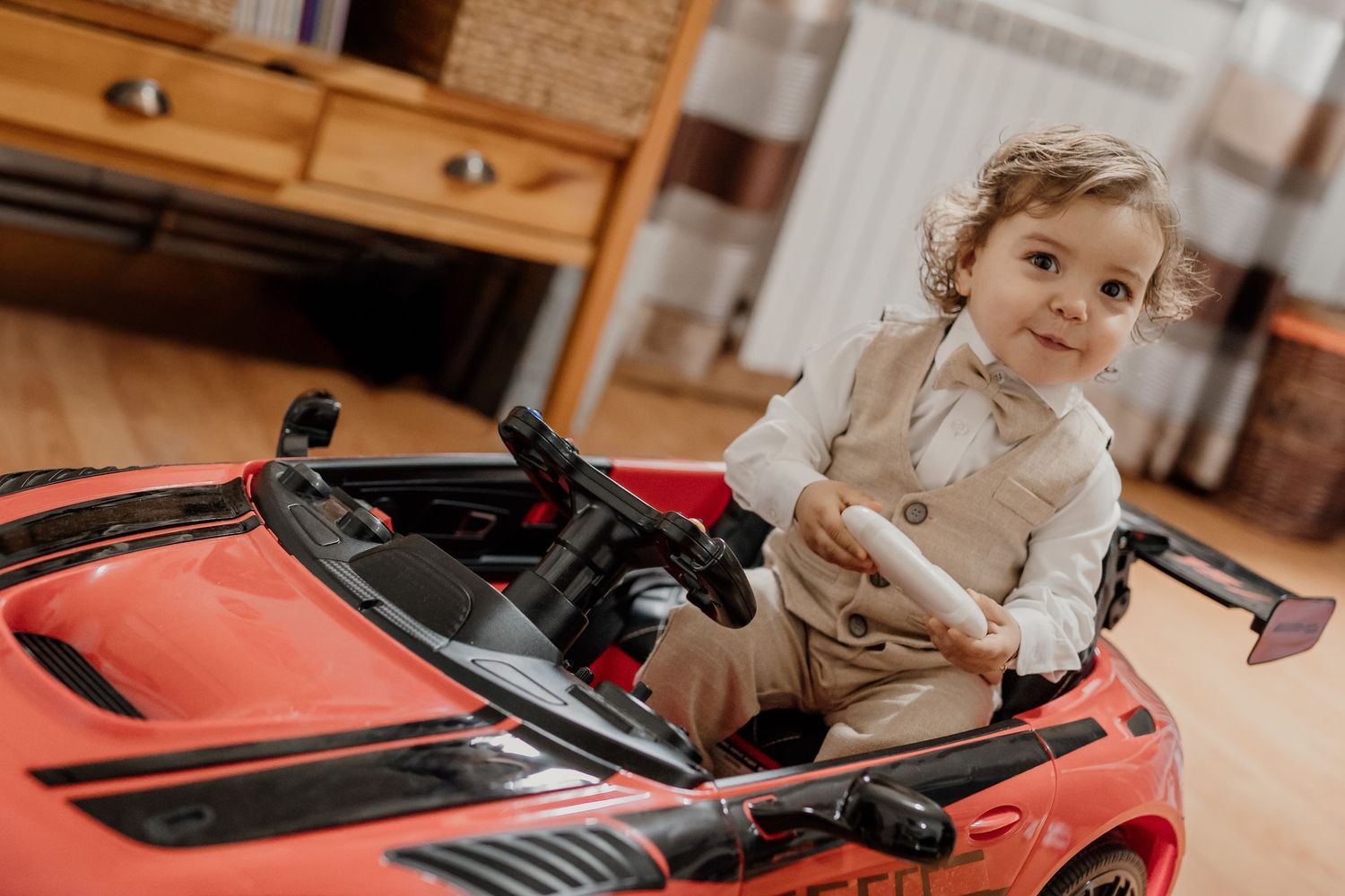 Toddler boy in white shirt sitting in red ride-on toy sports car on wooden floor indoors.