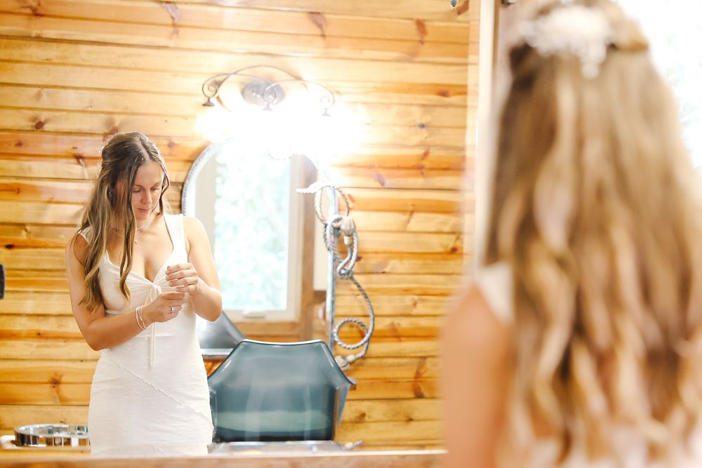 Bride in white dress applying perfume while looking in oval mirror inside rustic wood-paneled bridal suite.
