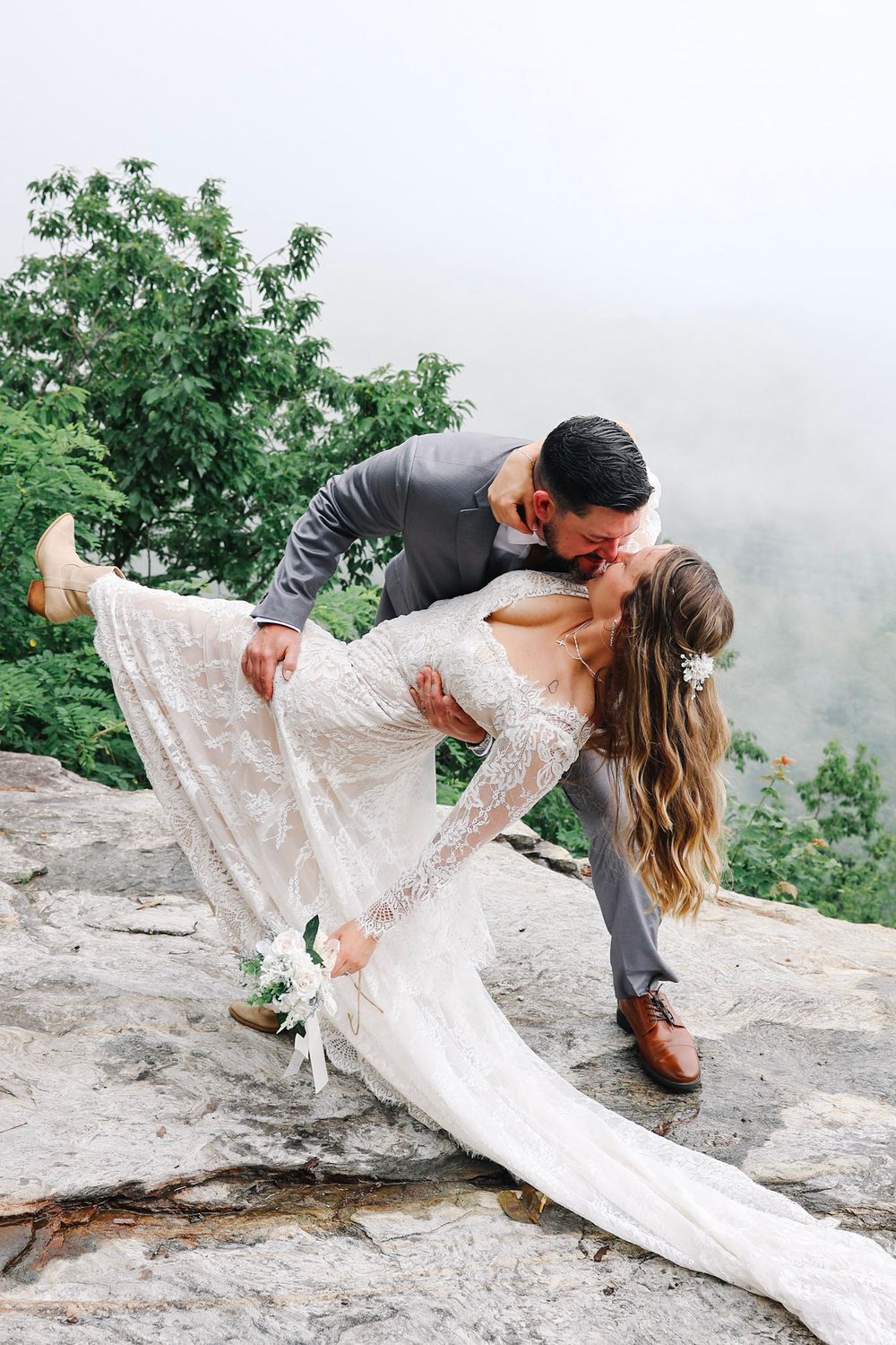 Groom dips bride for a romantic kiss on a rocky overlook with misty green mountains in the background.