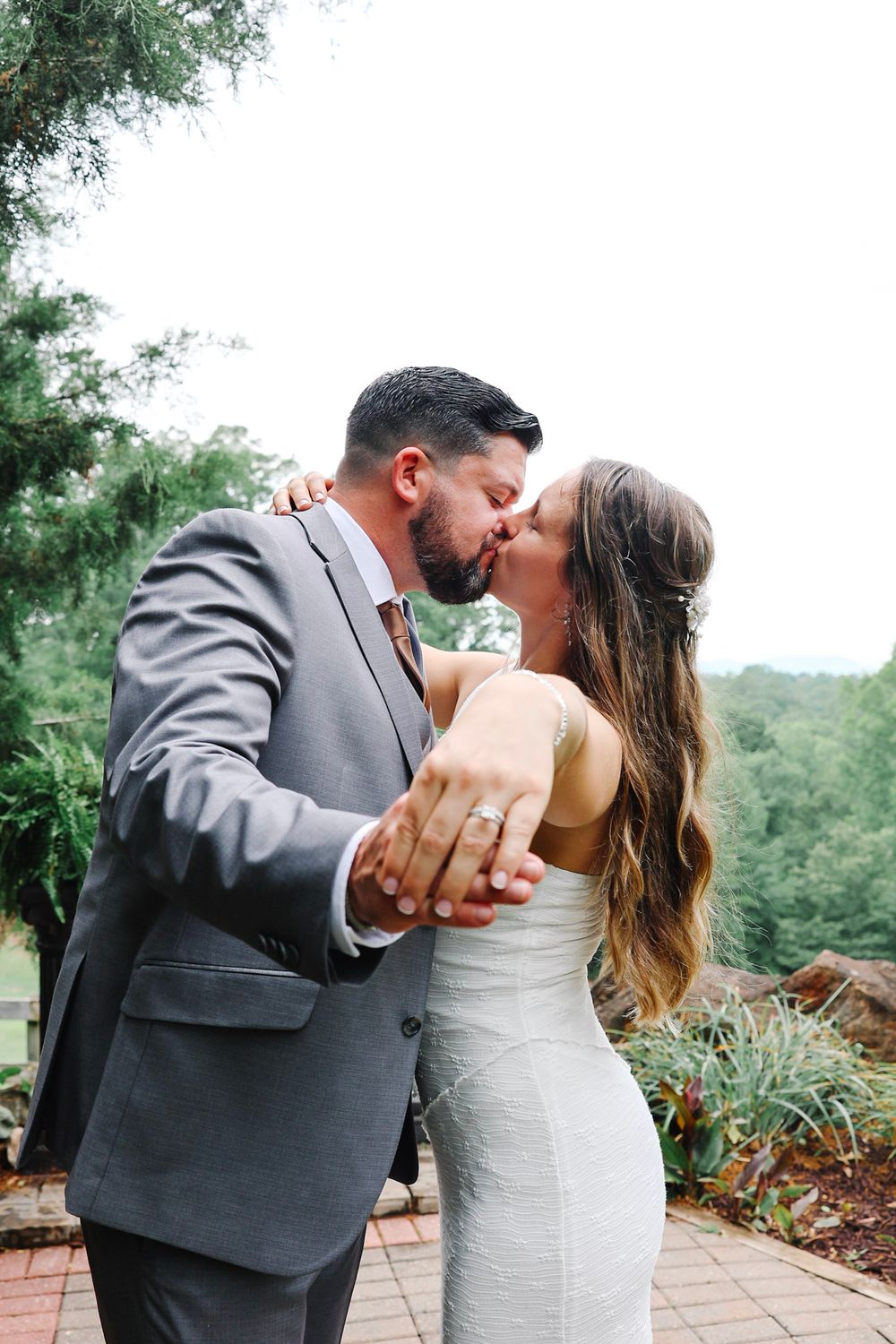 Newlyweds share a kiss while showing off wedding rings, bride in white dress, groom in gray suit, outdoor garden setting.