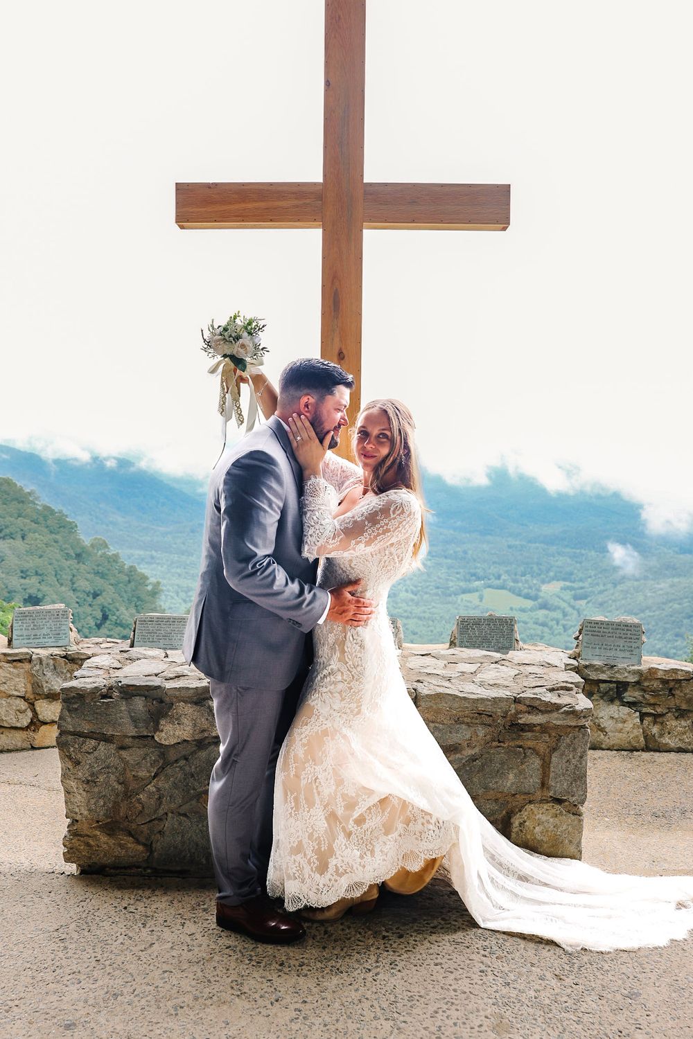 Bride and groom share a tender kiss beneath a wooden cross with scenic mountain views at their wedding.