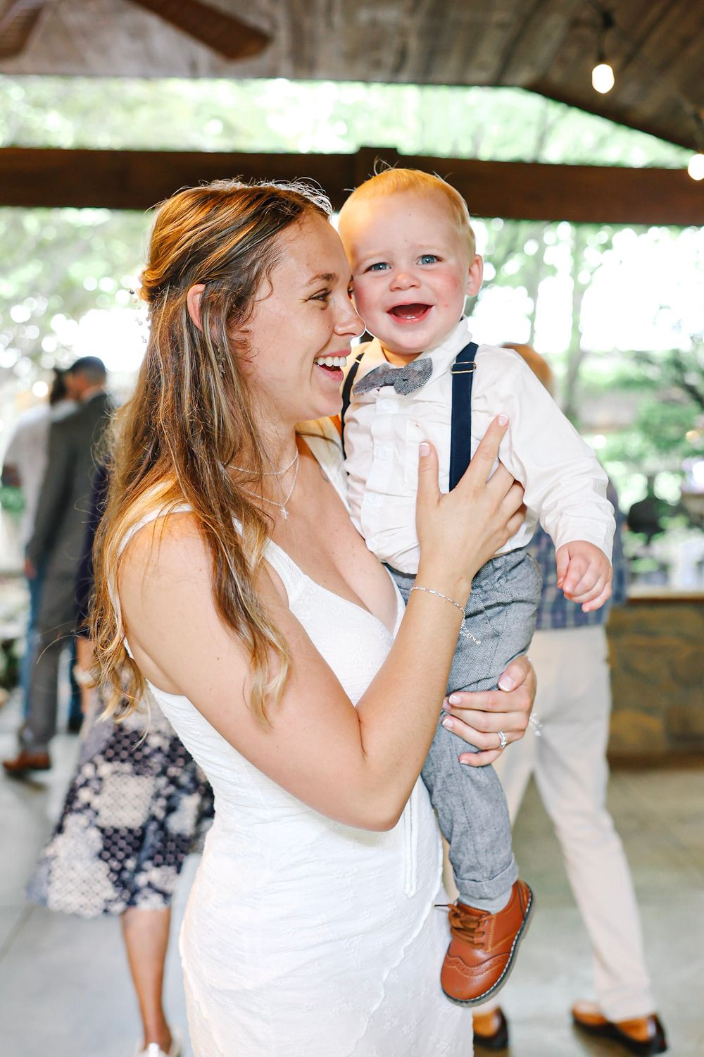 A joyful bride in a white dress holds a laughing baby boy in suspenders and bow tie at a wedding reception.