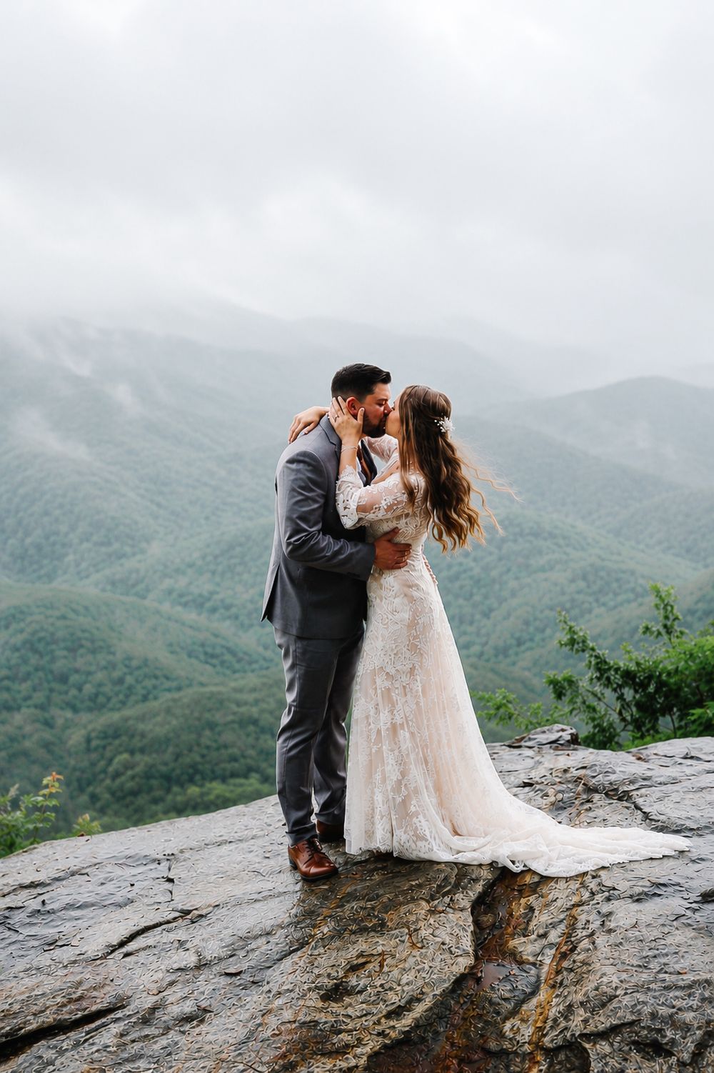 Bride and groom kissing on rocky mountain overlook with misty green mountain range backdrop.